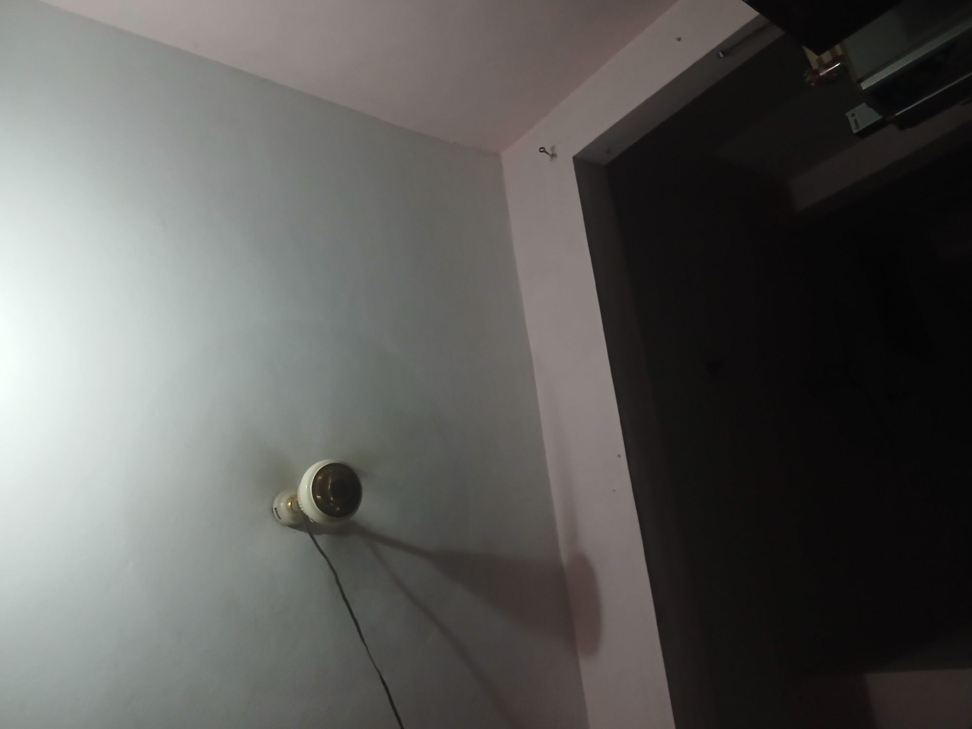 The image shows a ceiling with a light fixture and a hook on the wall. The ceiling is painted white and appears to be the interior of a room. The light fixture is white with a brass-colored base and a dark bulb. A black wire is connected to the light fixture and extends to the left. The wall below the ceiling is painted a light pink color. On the pink wall, there is a small, metal hook. The rest of the image is dark, suggesting the room is dimly lit, except for the light from the fixture. This scene is located in Peshawar City Tehsil, Pakistan.