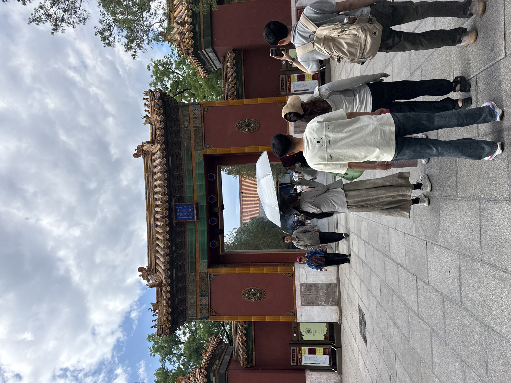 A group of tourists stand in a courtyard in front of an ornate, traditional Chinese gate in Dongcheng District, China. The gate is painted red with yellow accents and features intricate roof decorations and a blue plaque with Chinese characters. The ground is paved with gray tiles. Several people are visible, including a couple walking towards the gate, one holding an umbrella, and a group of younger individuals facing away from the camera, some holding phones. The sky is partly cloudy.