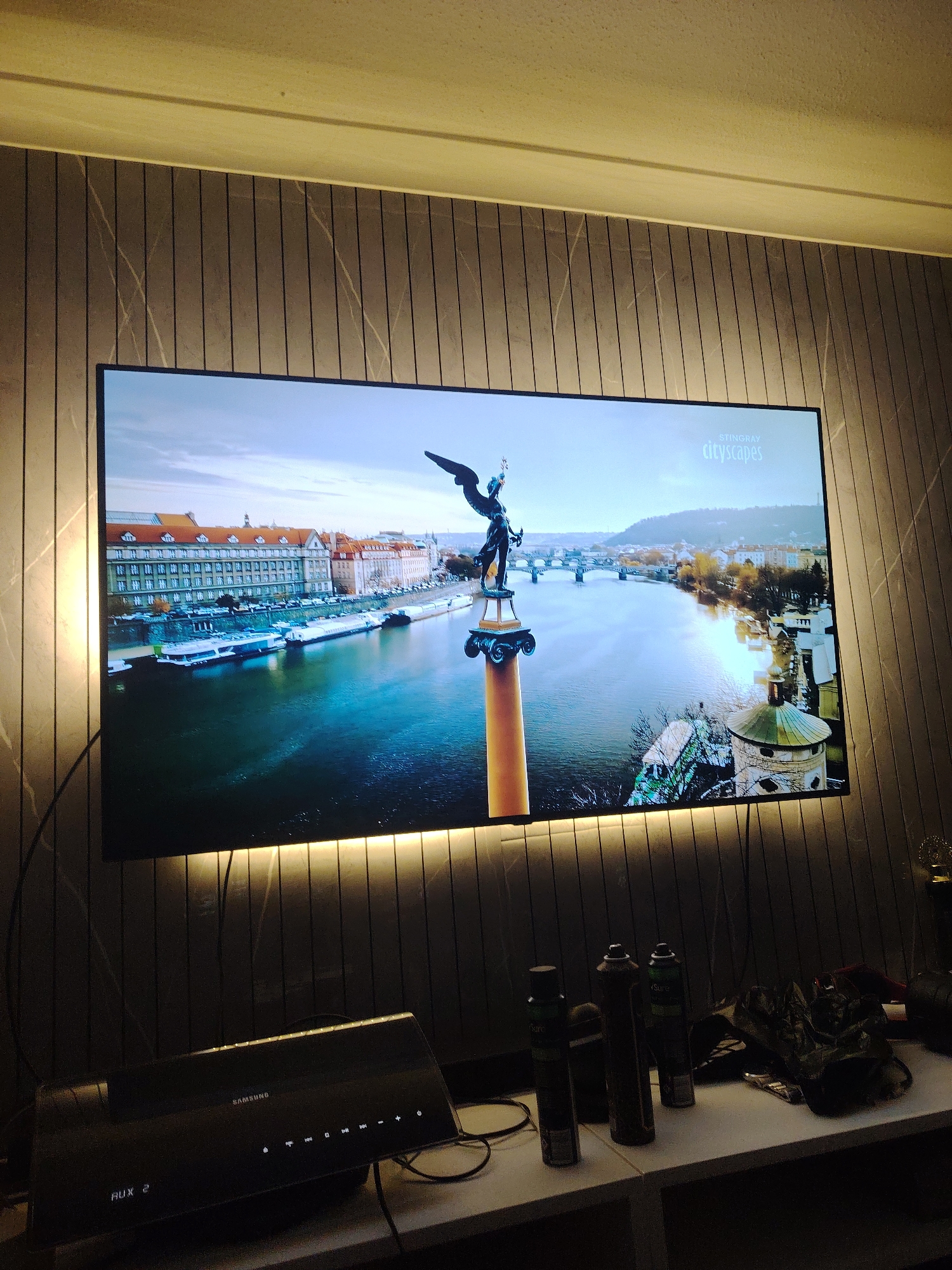 This image captures a domestic interior, located in Huntingdonshire, United Kingdom. A large, wall-mounted television screen dominates the center, displaying an aerial cityscape. The city on screen features a wide river flowing through it, spanned by multiple bridges in the background. In the immediate foreground of the screen, a dark, winged statue on a tall, orange-colored classical pedestal with a circular base is prominently displayed, its left arm raised. Buildings with numerous windows and pitched, reddish-orange roofs line the riverbanks, where several long, white tour boats are docked. A green-domed circular tower is visible on the right side of the river. The sky above the cityscape is overcast. A white "STINGRAY cityscapes" logo is visible in the upper right corner of the television screen.
The television is set against a wall featuring dark vertical panels with subtle, irregular white vein-like patterns. Warm yellow LED strip lights illuminate the wall both above and below the television. Above the paneled wall, a light-colored molding or trim runs horizontally, beneath a textured, light beige ceiling.
Below the television, a white, horizontally oriented shelf holds several items. On the left, a black Samsung soundbar or media device is visible, displaying "AUX 2" in illuminated white text on its left side, and a series of illuminated control buttons (power, volume, media controls) on its right. Black cables are visible trailing from behind the television and across the shelf, connecting to the Samsung device. To the right of the Samsung device, three dark spray cans stand upright, with "Sure" branding partially discernible on two of them. Behind the cans, a crumpled dark object, possibly a bag, rests on the shelf. A golden, ornate decorative item or trophy is partially visible on the far right edge of the shelf. The room is generally dimly lit, with the television and LED strips providing the primary illumination. No individuals are present in the scene.