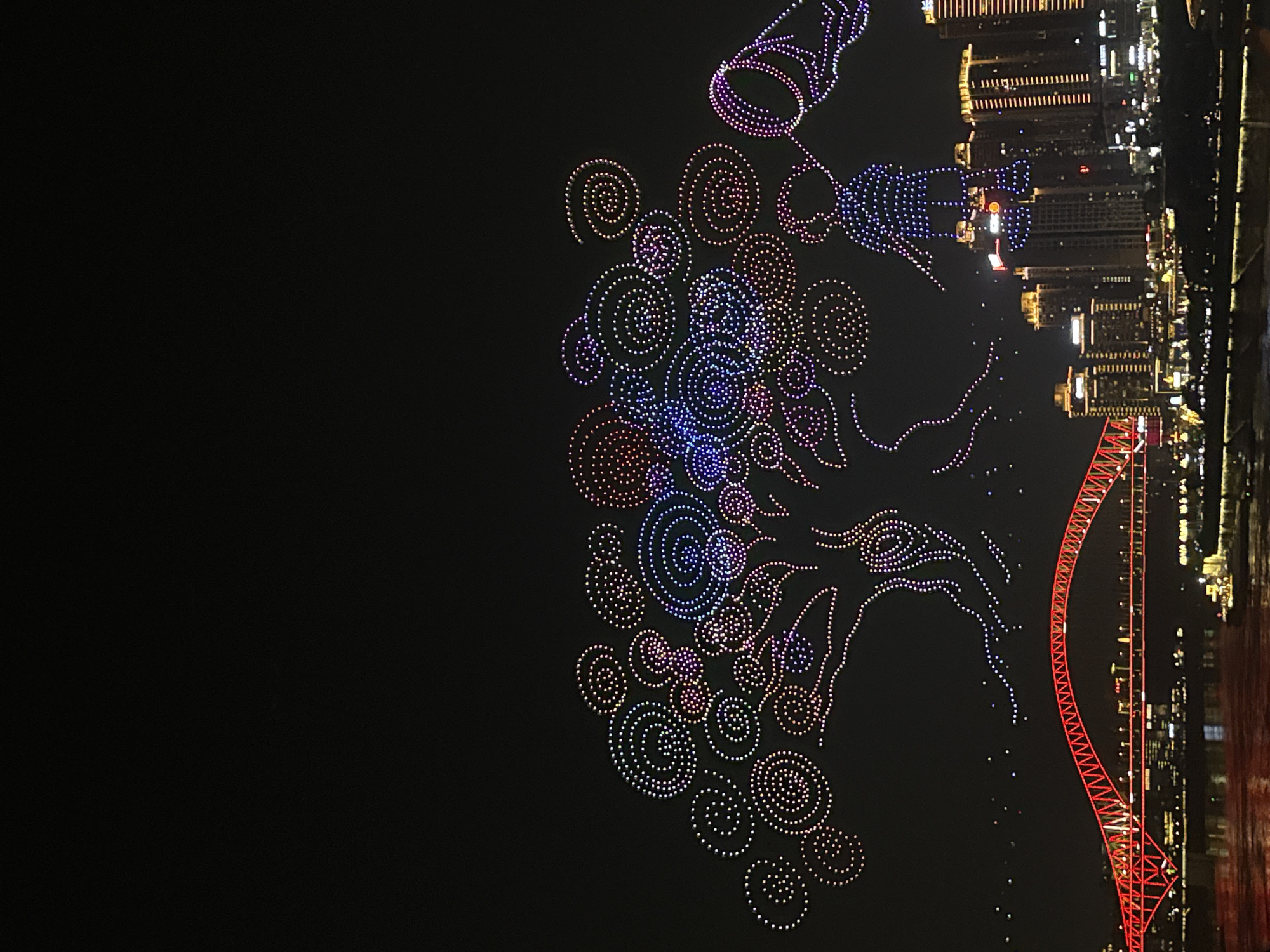 A nighttime aerial drone light show depicts a giant tree formed by thousands of illuminated dots, with swirling patterns and a human-like figure made of lights to the right of the tree. Below the drone display, a city skyline is visible with illuminated buildings. A large, red, arched bridge spans across a river in the foreground. The scene appears to be taking place in the Yuzhong District of Chongqing, China, given the distinctive architecture of the city and the bridge.