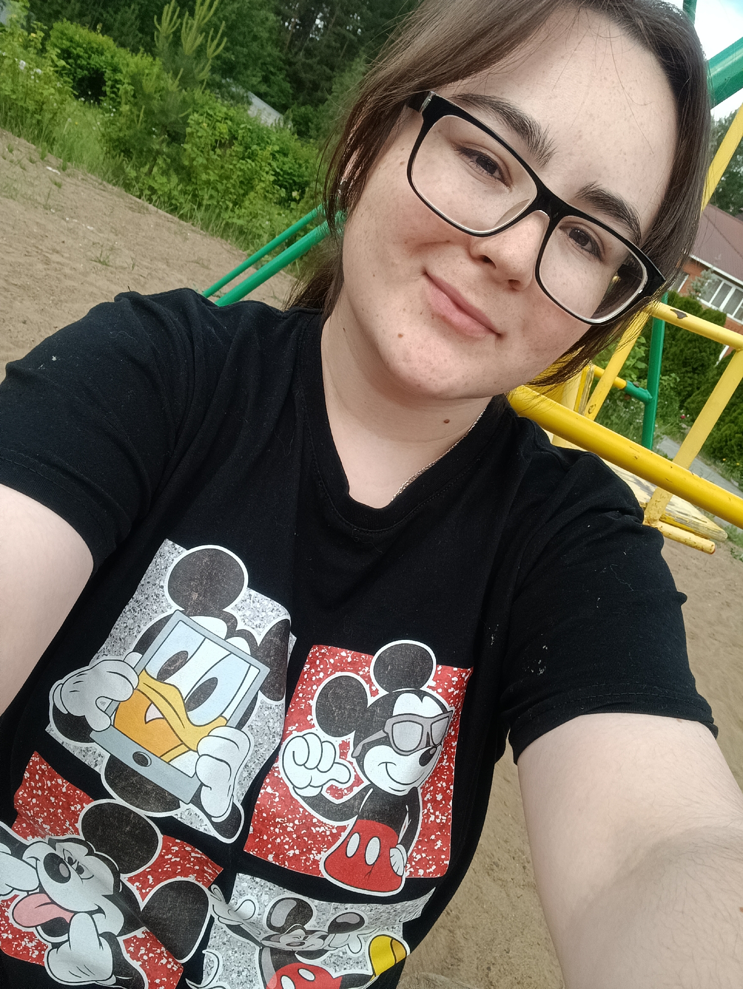 A young woman wearing glasses and a black t-shirt with a Mickey Mouse and Donald Duck design takes a selfie. The background consists of a sandy area, a green and yellow metal structure, likely part of a playground, and some greenery including trees. A building roof is partially visible in the upper right corner. The photo was taken in the city of Chaikovsky, Russia.