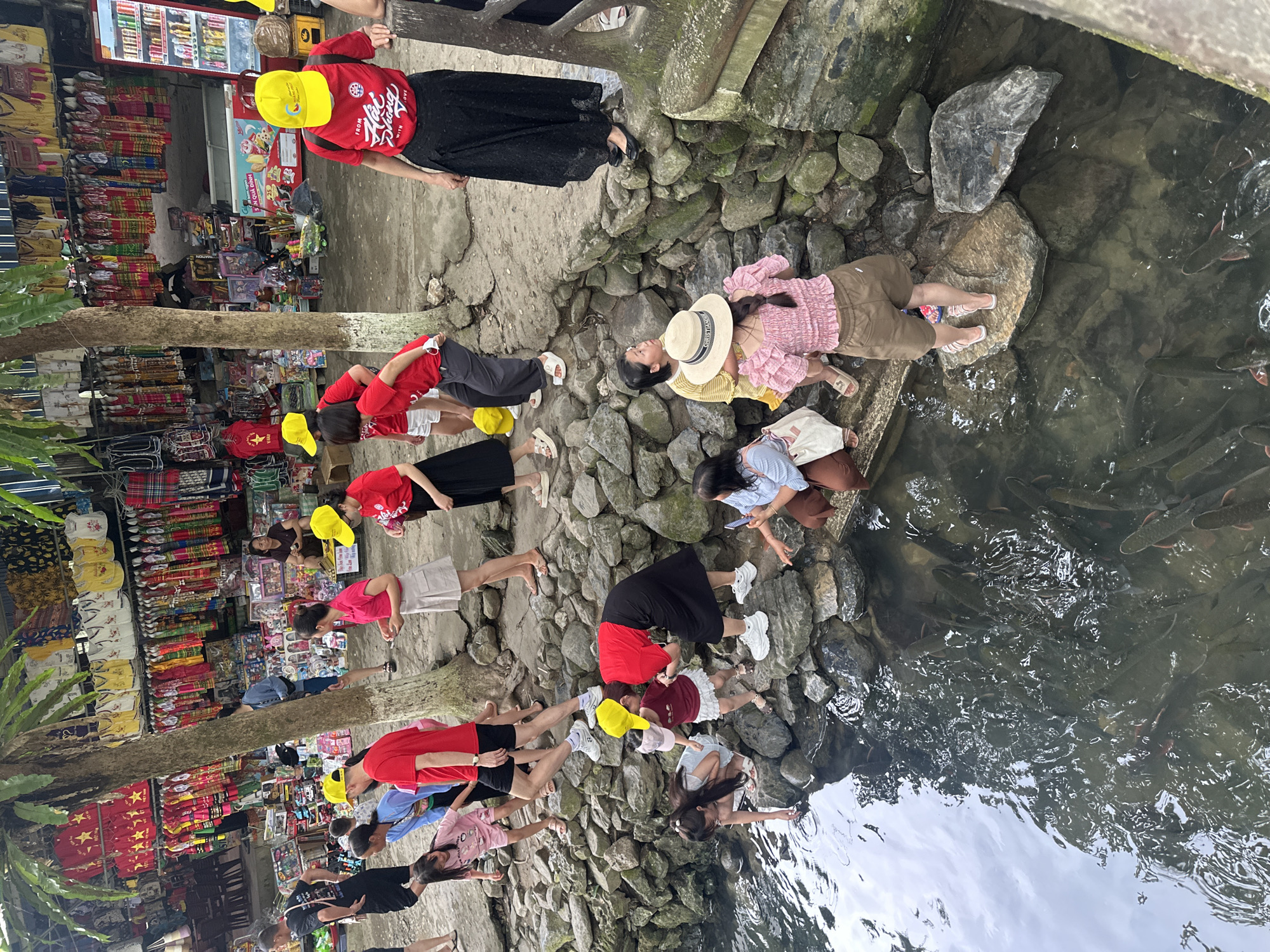 The image shows a group of people, including adults and children, gathered by a shallow body of water filled with fish. The setting appears to be an outdoor market or tourist attraction, with stalls selling clothing and other goods in the background. Several individuals are wearing yellow hats, and some are interacting with the water, possibly feeding the fish. The ground is rocky, and lush green trees provide shade. The overall atmosphere suggests a casual and lively environment. The location is identified as Cẩm Thạch Commune, Vietnam.
