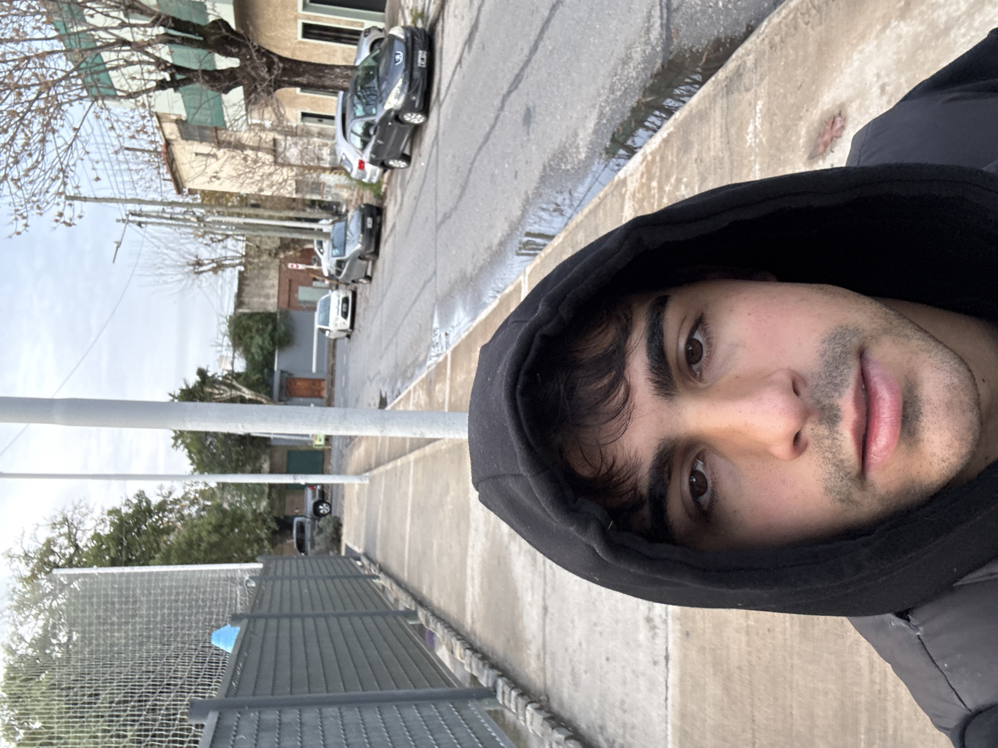 The image is a selfie taken from a low angle on a street in Avellaneda, Argentina. The subject, a young man with dark hair, is wearing a black hooded jacket and the hood is up. He is looking directly at the camera. In the background, there is a sidewalk with a grey fence on the left and a street with several parked cars. The street appears wet, possibly from recent rain. Buildings line the right side of the street. Overhead power lines are visible. The sky is overcast.
