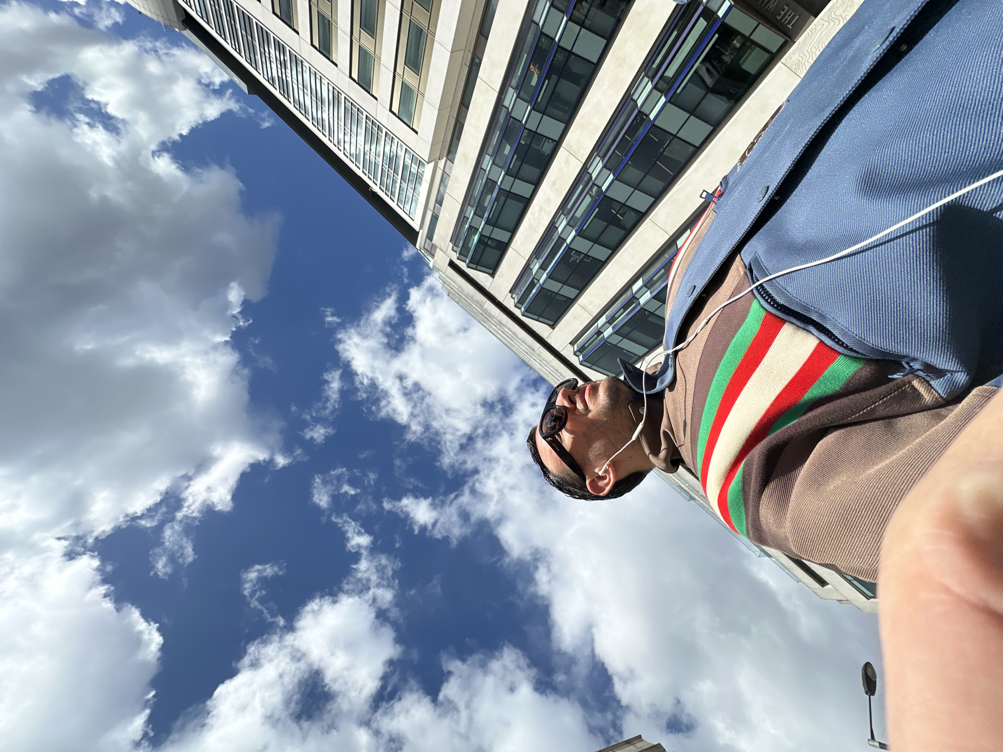 A low-angle outdoor image depicts a male individual in the foreground against a backdrop of a modern building and a partly cloudy blue sky. The scene is located in the City of London, United Kingdom, specifically outside The Walbrook building at 25 Walbrook, identifiable by the visible text "THE WAL" on the structure.
The subject, positioned centrally and looking upwards and to the right, wears dark sunglasses and white wired earbuds. He is dressed in a brown collared top featuring a distinctive red, green, and white horizontal stripe pattern across the chest and shoulder, partially covered by a blue outer garment. A blurred digit, likely a thumb or finger, occupies the bottom-left corner of the frame.
To the right, a tall, multi-story modern office building with a light-colored facade and numerous reflective windows rises into the frame. Some windows exhibit a gold tint while others reflect the blue sky. On the left, a less distinct building and a black street lamp are partially visible. The sky features substantial white cumulus clouds against a clear blue background, indicating bright daytime conditions.
