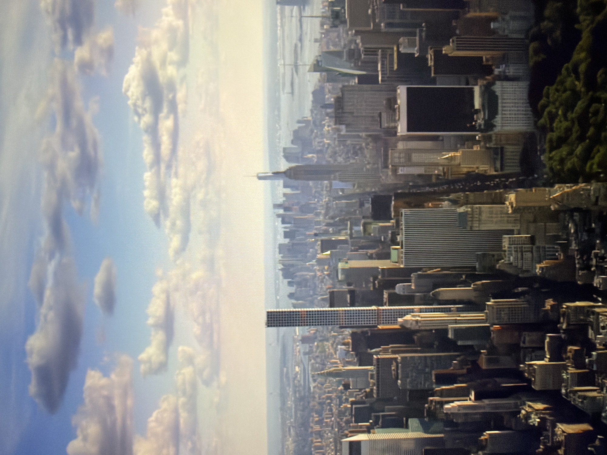 The image is an aerial view of a cityscape bathed in sunlight under a partly cloudy sky. Dominating the foreground is a dense collection of skyscrapers of varying heights and architectural styles. A very tall, slender skyscraper with a checkered facade stands prominently in the mid-ground. Further into the background, the cityscape extends to the horizon, where a body of water can be seen, possibly a river or harbor, with more distant landmasses and structures. The scene depicts an urban environment, likely a major metropolitan area, characterized by its extensive development and high-rise buildings. The location is not Kyiv, Ukraine; it is a view of New York City.