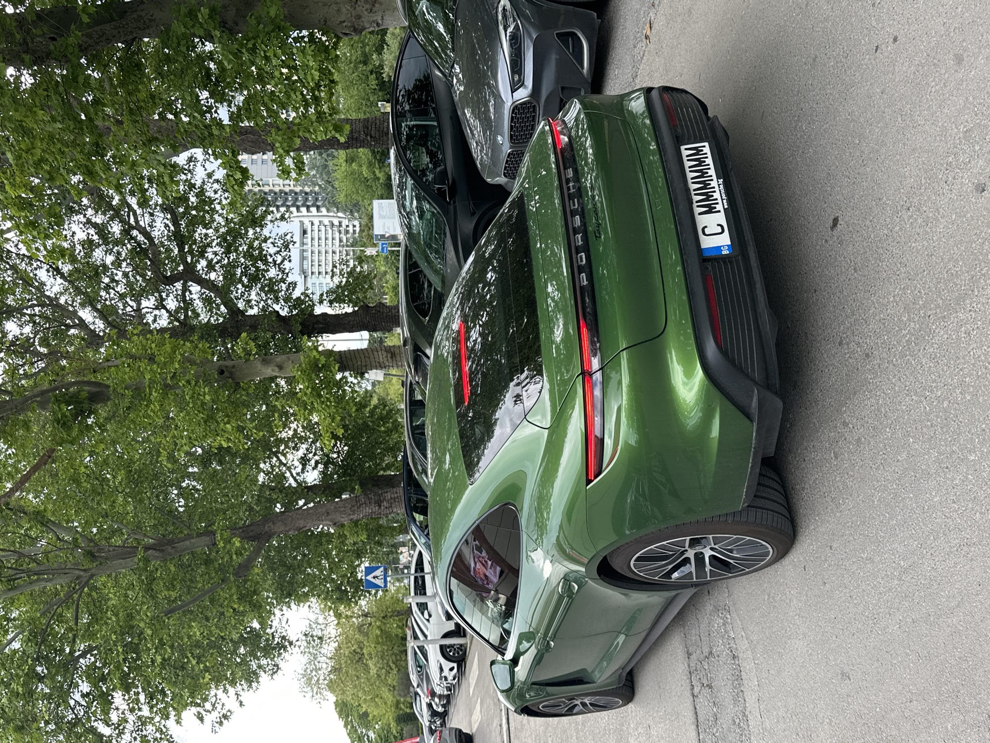 The image depicts a green Porsche Taycan parked on a street in Varna, Bulgaria. The car has a license plate reading "C MMMMMMM" with the Bulgarian flag. Several other cars are parked behind the Porsche. The background includes trees and a high-rise building. The location appears to be an urban setting, with the cars parked along a road with sidewalk.