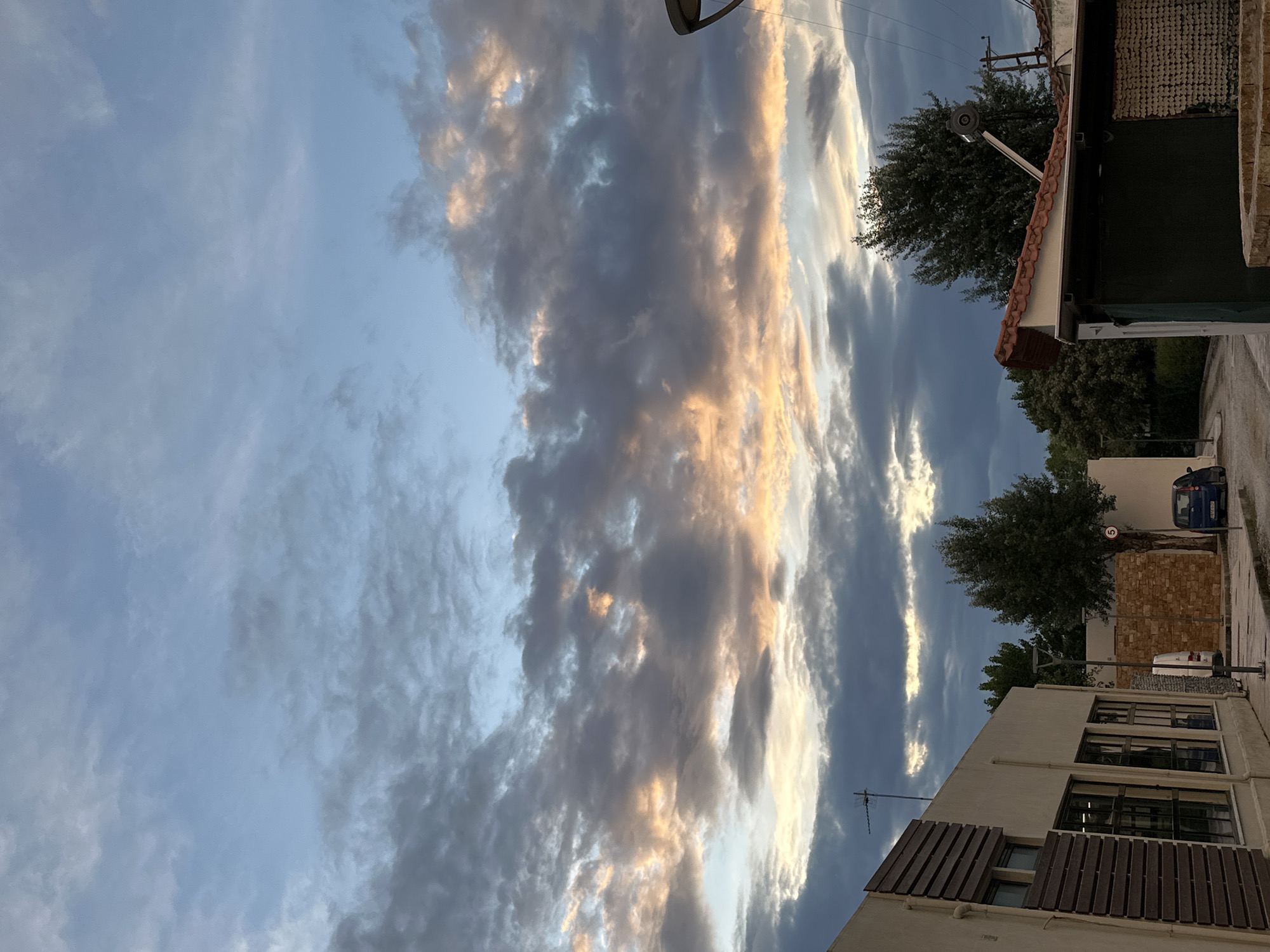 The image, taken in Ofrynio Beach, Greece, shows a view from street level looking upwards, primarily showcasing the sky and the tops of adjacent buildings. The sky is filled with a dramatic cloud formation, a mix of dark, voluminous clouds interspersed with areas illuminated by the setting sun, casting a golden hue on the cloud edges.
At the bottom of the frame, sections of two buildings are visible. The building on the left side features a white facade with dark window frames and brown wooden shutters. An antenna is mounted on the roof. The building on the right is more fragmented in view, but it reveals a terracotta tiled roof, a security light, and an adjacent building covered in dark netting.
Further down the street, a section of brick wall, a small tree, and a parked blue car are visible, partially obscured by the buildings. The scene is generally still and quiet, with the primary visual interest being the dynamic sky.