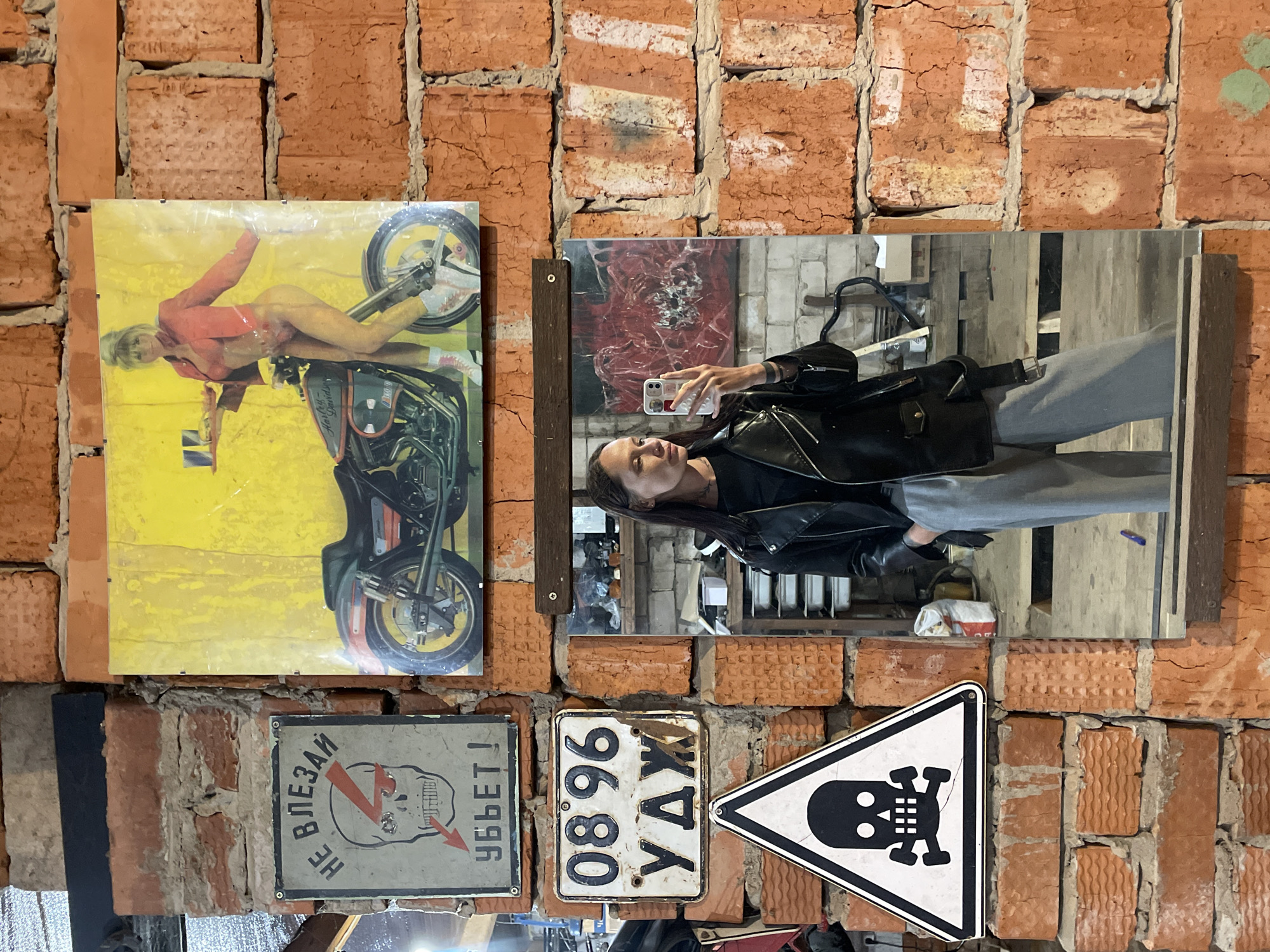 The image shows a brick wall decorated with various items. These include a poster of a woman on a motorcycle, a mirror reflecting a woman taking a selfie, two Russian-language warning signs, and a triangular sign with a skull and crossbones. The location is Oktyabr'skii, Russia, inferred from the Russian writing on the license plate.