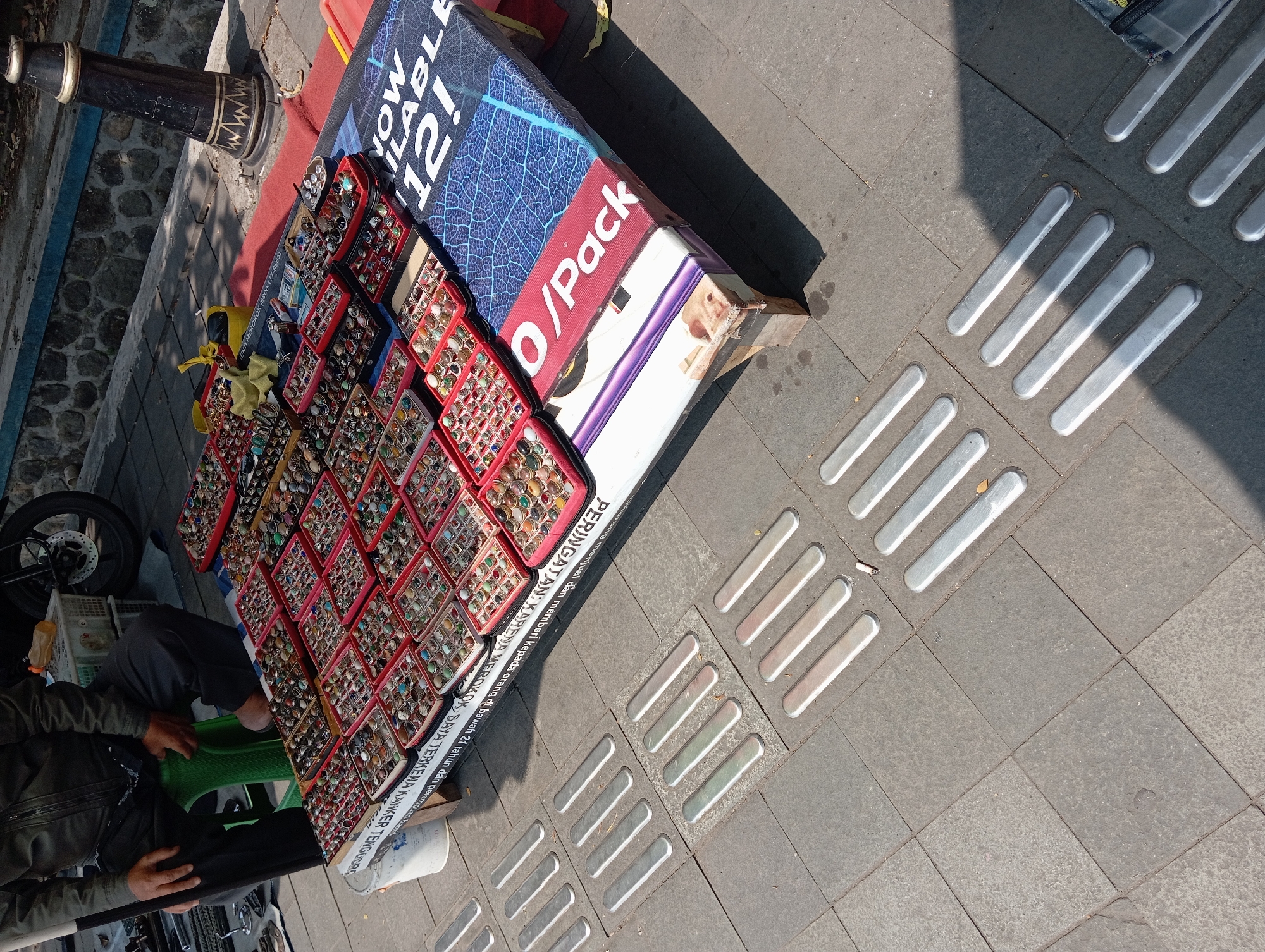 A vendor is sitting on a green stool to the left, selling rings displayed in trays on a table. The table has a colorful, patterned cover. The background shows a stone wall and a portion of a motorcycle. The foreground consists of grey paving stones with raised tactile paving indicators. The scene appears to be outdoors, likely a market or street. The overall impression is of a typical street-side retail setup.
