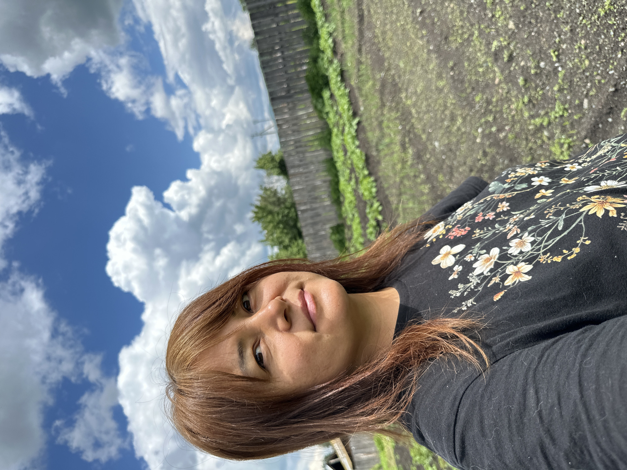 A medium shot captures a woman with reddish-brown hair and bangs looking directly at the camera. She is wearing a black t-shirt with a floral print. The background reveals a cultivated garden with rows of green plants, a rustic wooden fence, and a bright blue sky filled with fluffy white clouds. A portion of a wooden structure is visible on the far left. The scene appears to be outdoors in a rural or suburban setting.
