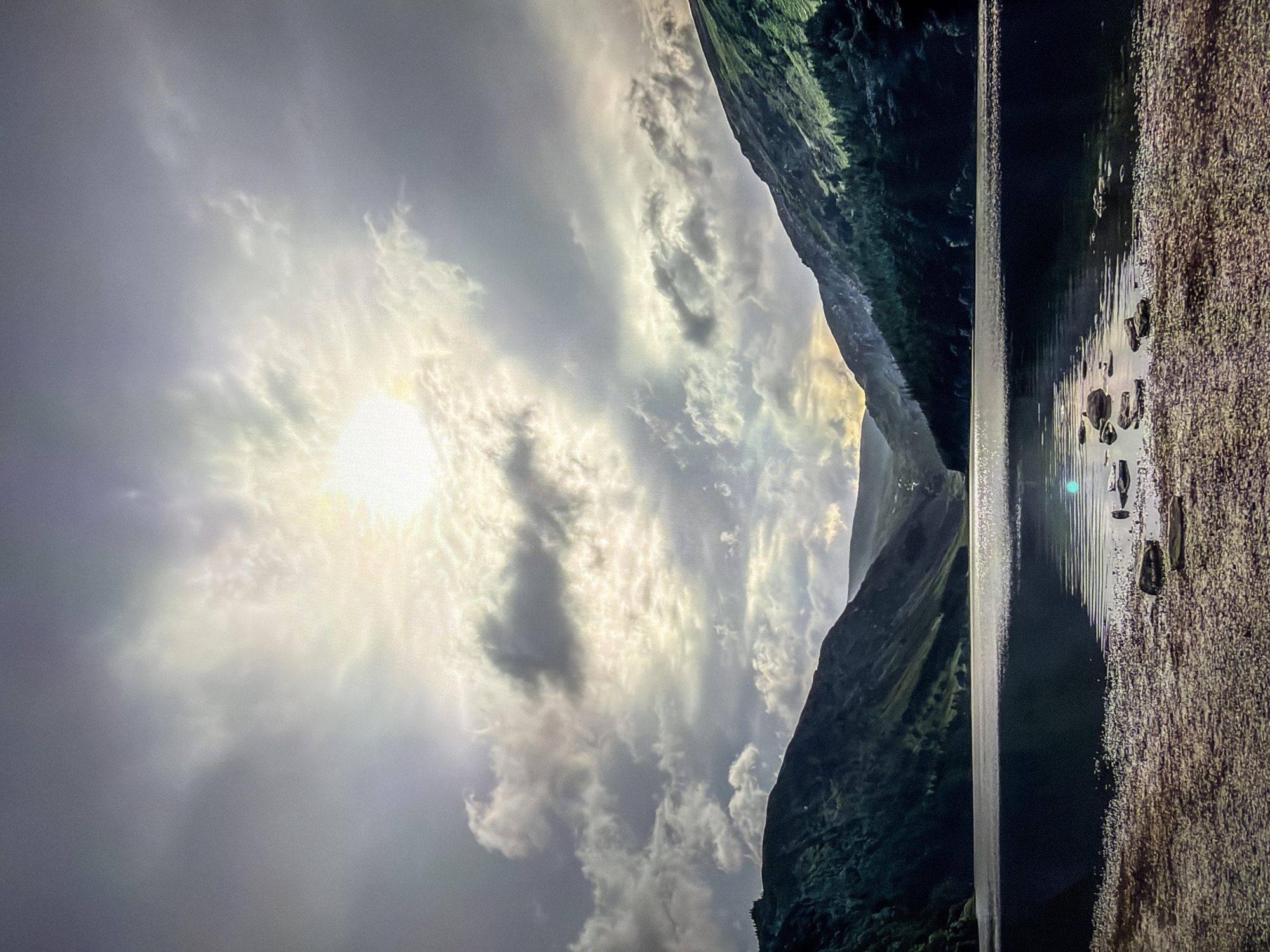 This media file presents a striking natural landscape, likely a lake or fjord nestled between mountains under a dramatic sky. The image is dominated by a bright sun, positioned towards the upper left, which casts diffused light through and around an expanse of scattered clouds. The sky transitions from a luminous white and pale yellow around the sun to darker grey and bluish tones, with distinct cloud formations showing texture.
Dark, verdant mountains or steep hills frame the body of water. The mountain on the left appears more distant and is partially shrouded in atmospheric haze, while the closer mountain on the right exhibits rugged, dark green foliage and rocky details.
Below, a tranquil body of water stretches across the lower part of the frame, acting as a mirror to the sky and sunlight. Bright, elongated reflections and ripples shimmer on its surface, contrasting with the deeper, darker sections of the water. To the far right, a textured shoreline or bank, composed of pebbles or small rocks, meets the water, also illuminated by the sun's rays.
The lighting suggests either early morning or late afternoon, with the low sun creating strong backlighting, dramatic highlights, and deep shadows across the landscape. The overall mood is one of majestic serenity and natural beauty. No people or visible text are present in the image. The scene is consistent with the natural landscapes found in Ros Na Rí, Ireland.