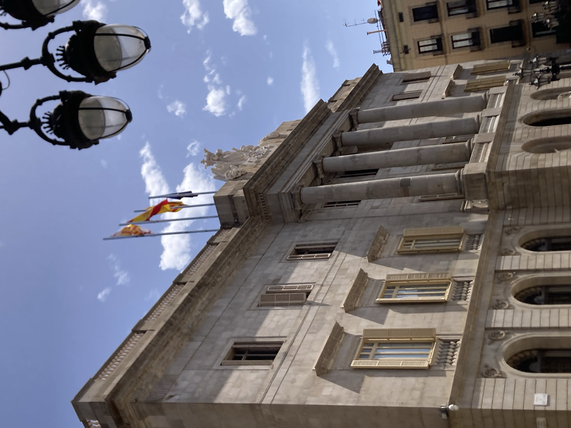 The image shows the exterior of a grand, ornate building, likely a government or public building, with a neoclassical architectural style. The building is constructed of stone and features prominent columns, decorative sculptures at the top, and numerous windows. Two flags, one appearing to be the Spanish flag and another a regional flag, are visible on flagpoles. The perspective is from a low angle, looking up towards the sky, which is a bright blue with scattered white clouds, suggesting a sunny day. In the foreground, the top of a street lamp with multiple globes is visible, indicating an urban setting. Another building, taller and more modern with visible antennas, is seen behind the main building. The lighting suggests it is daytime. The overall impression is one of civic grandeur and historical significance.