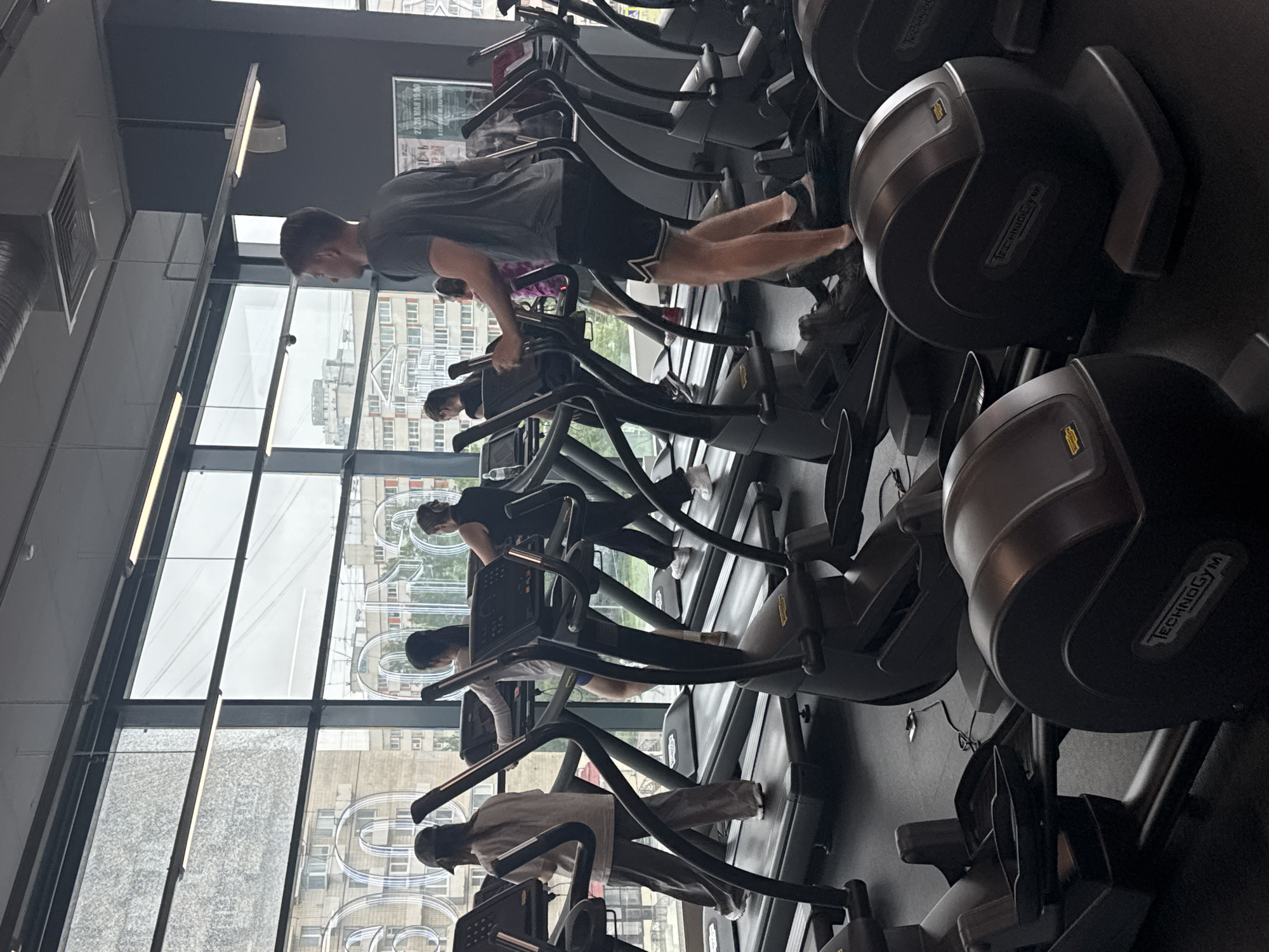 A row of several elliptical machines are occupied by people working out, located in a gym in Saint Petersburg, Russia. The machines are black with the brand "Technogym" visible. The people are wearing athletic clothes and facing toward a large window with views of buildings and the city skyline.