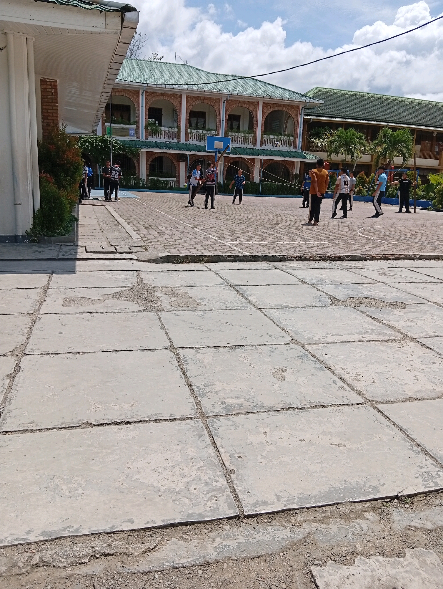 The image depicts an outdoor scene at what appears to be a school courtyard in the City of Gunungsitoli, Indonesia. The foreground is dominated by large, worn concrete slabs, some of which are cracked and have rough edges. Beyond this, a paved area with brick-like patterns serves as a basketball court. Several young men in athletic wear are engaged in activities on the court. In the background, a multi-story building with arched windows and a green tiled roof is visible. Palm trees and other greenery are interspersed around the building. The sky is partly cloudy with scattered white clouds. The overall impression is of a school campus on a bright day.