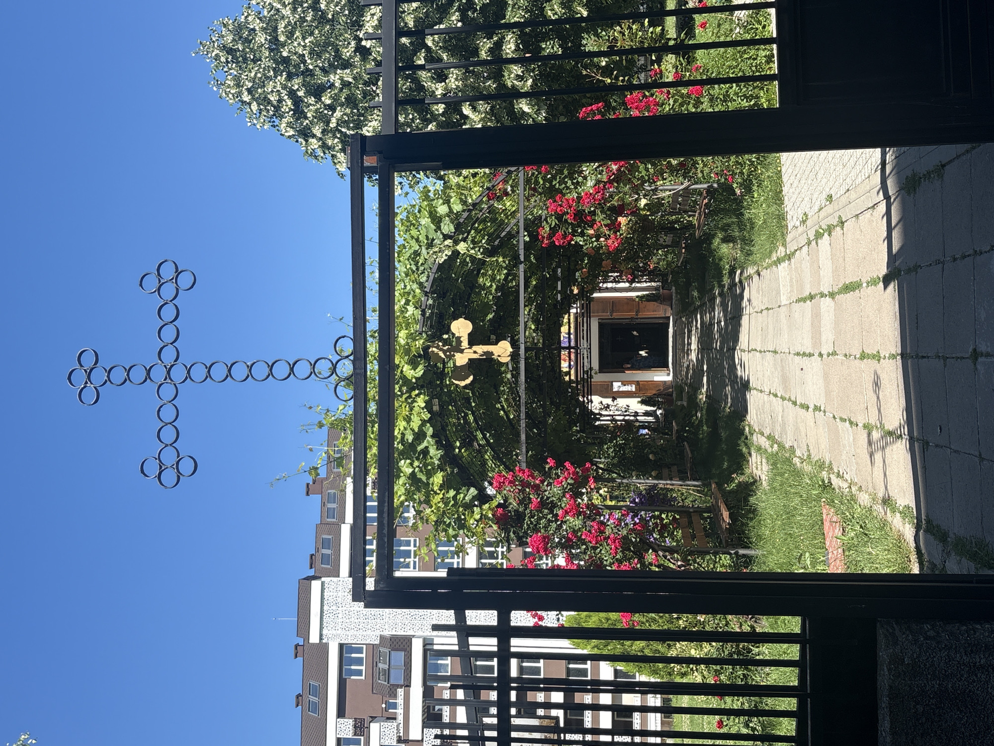 The image shows a view through an open black metal gate into a courtyard or garden area. Above the gate, a large, ornate cross made of interconnected circles is silhouetted against a clear blue sky. The gate opens onto a paved walkway leading to an arched walkway covered with green foliage and red roses. A golden cross is visible hanging from the archway. Beyond the archway, the entrance to a building, possibly a church, is visible with its doors open. In the background, to the left, are modern apartment buildings with brown roofs. To the right, a large, leafy tree with white blossoms is visible. The ground is covered with grass and the walkway is made of rectangular paving stones. Sunlight casts shadows across the walkway.