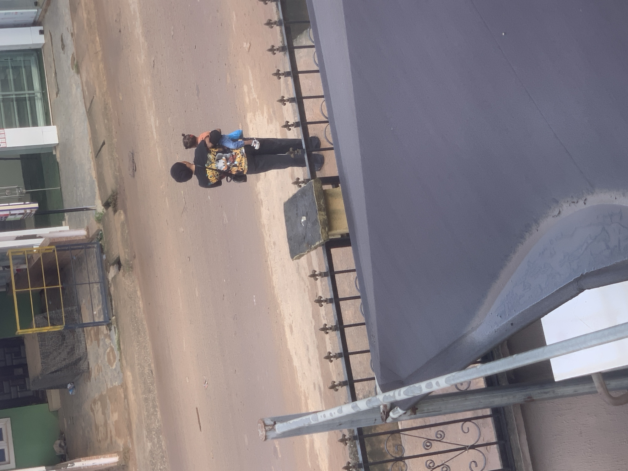 In the city of Shagamu, Nigeria, a person stands holding a child in their arms. The individual is wearing a black t-shirt with a graphic design and black pants. The child, facing away from the camera, is wearing an orange top and blue pants. They are standing on a dirt road, with a black metal fence in the foreground and buildings in the background. The overall scene suggests a residential or commercial street environment.