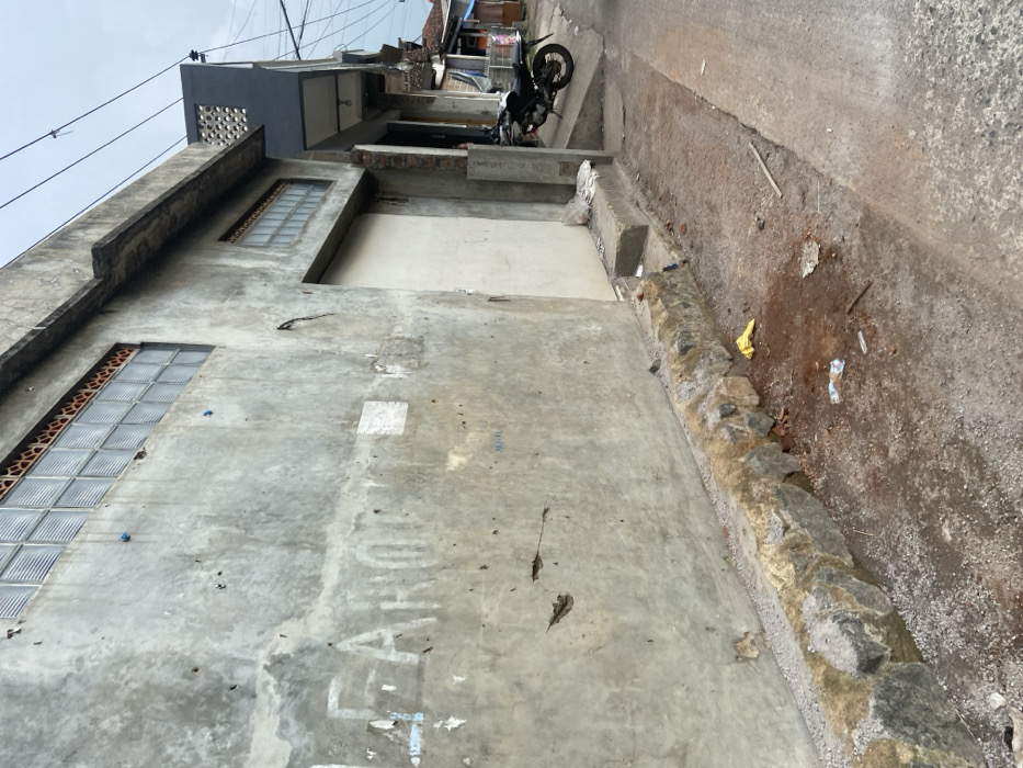 The image shows the side of a concrete building with a large, white garage door. The building has a row of windows made of small glass squares and a concrete foundation. The building has some damage to the exterior, including cracks and missing sections of concrete. There is a row of large rocks along the base of the building and a small, yellow plastic bag lying on the ground in front of it. The building is located on a narrow, paved street. There is a motorcycle parked on the street to the right of the building. The location is a street in the city of Margaasih, Indonesia, based on coordinates (latitude: -6.957, longitude: 107.549).