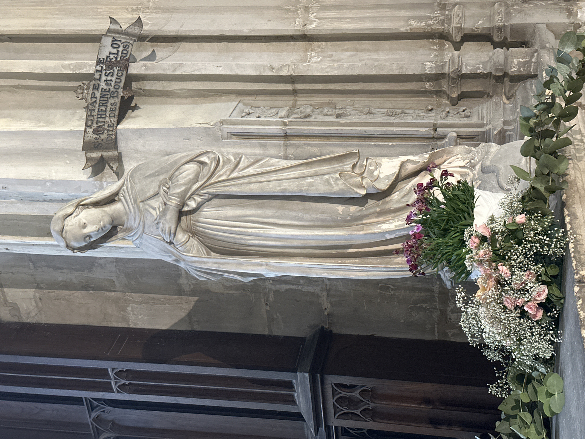 The image shows a statue of the Virgin Mary in a chapel dedicated to Saint Catherine and Saint Eloi, also known as the "Chapelle des Bouchards," located in Dieppe, France. The statue is made of white stone and depicts the Virgin Mary with her hands clasped at her chest. She is wearing a draped robe and veil. The statue is placed on a stone pedestal adorned with floral arrangements. The flowers include pink roses, small white baby's breath, and some purple orchids. Eucalyptus branches are also visible in the foreground. The background features architectural elements of the chapel, including stone columns and a metal sign with the chapel's dedication inscribed on it. The lighting in the scene is subdued, suggesting an indoor setting.