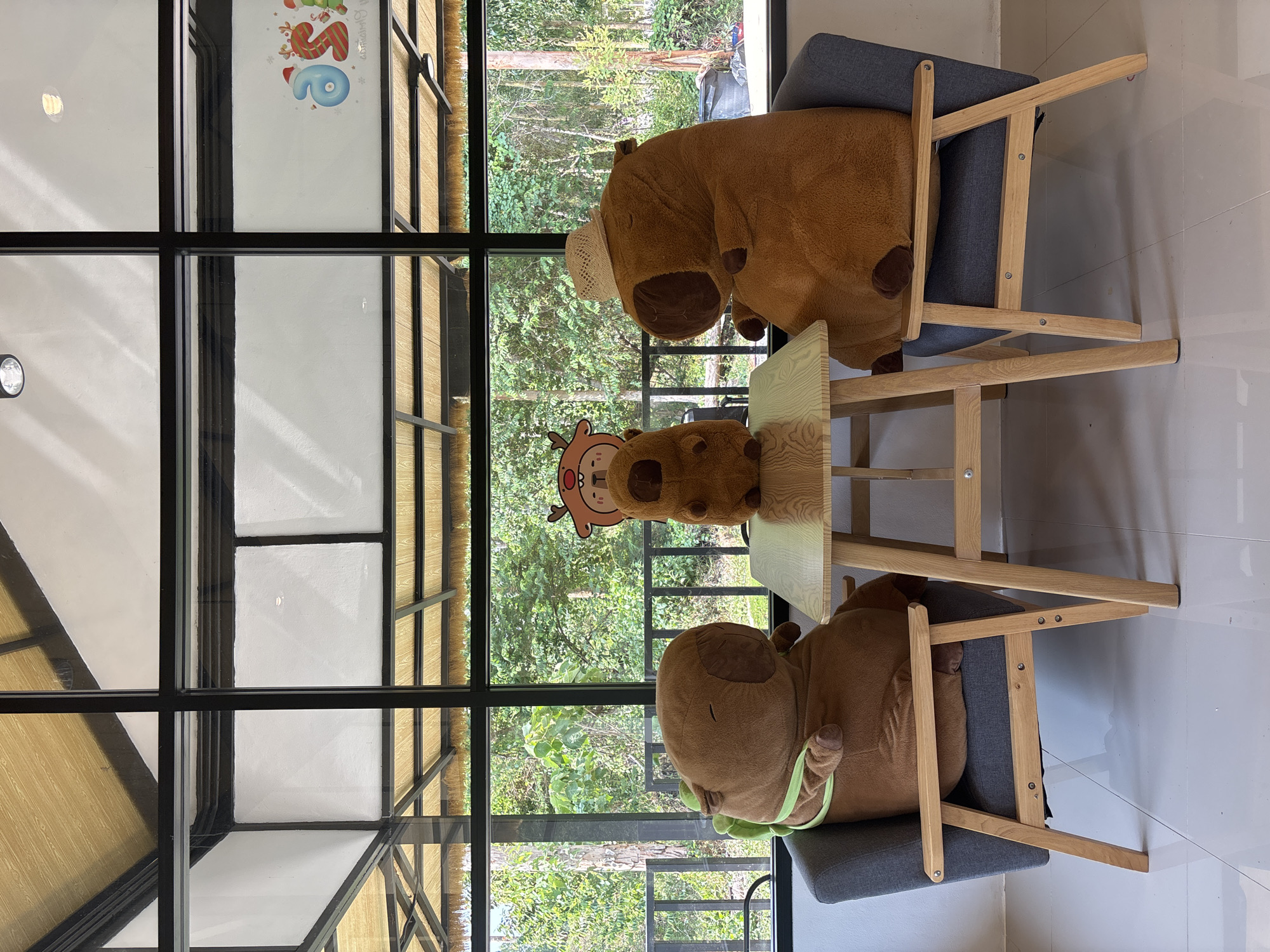 The image displays an indoor setting characterized by a large window overlooking a lush, green natural environment. Three plush toys are arranged around a small, light wooden table, creating a whimsical scene. Two larger, brown capybara-like plush toys are seated on grey upholstered chairs with wooden frames, positioned on opposite sides of the table. One of these larger plush toys wears a small straw hat, while the other has a light green strap around its body. A third, smaller, lighter brown plush, resembling a deer with antlers, is placed directly on the table.
Beyond the expansive window, which features black frames, a dense forest or thick vegetation is visible, indicating a natural and verdant outdoor setting. The bright natural light illuminating the trees suggests it is daytime with clear weather. Inside, the walls are a light, neutral color. A festive sticker or decoration is affixed to the windowpane in the upper left, featuring a cartoon reindeer head and colorful, striped letters that partially spell out "Happy S," hinting at a seasonal or celebratory theme. The overall atmosphere is playful and inviting, situated in a natural locale such as Choeng Thale, Thailand.