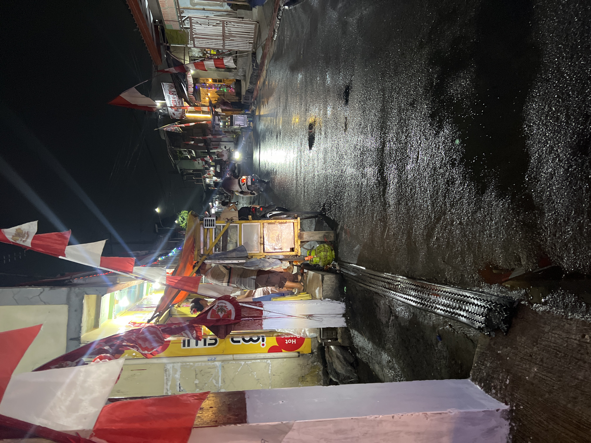 The image depicts a street scene at night in Citeureup, Indonesia, likely during a national holiday celebration given the prevalence of Indonesian flags. The wet asphalt street reflects the ambient light from shopfronts and streetlights, with several puddles visible. On the left, a building facade is partially visible, adorned with red and white Indonesian flags. In front of this building, there is a yellow sign. Next to it, a food cart is set up with a man and a woman attending to it, and a green gas cylinder is placed on the ground. Several more Indonesian flags are displayed along the street. Further down the street, motorcycles are visible, with one rider illuminated by headlights. Buildings and stalls line both sides of the street, with awnings and illuminated signs indicating commercial activity. The overall atmosphere is one of a bustling night market or street with a festive decoration.