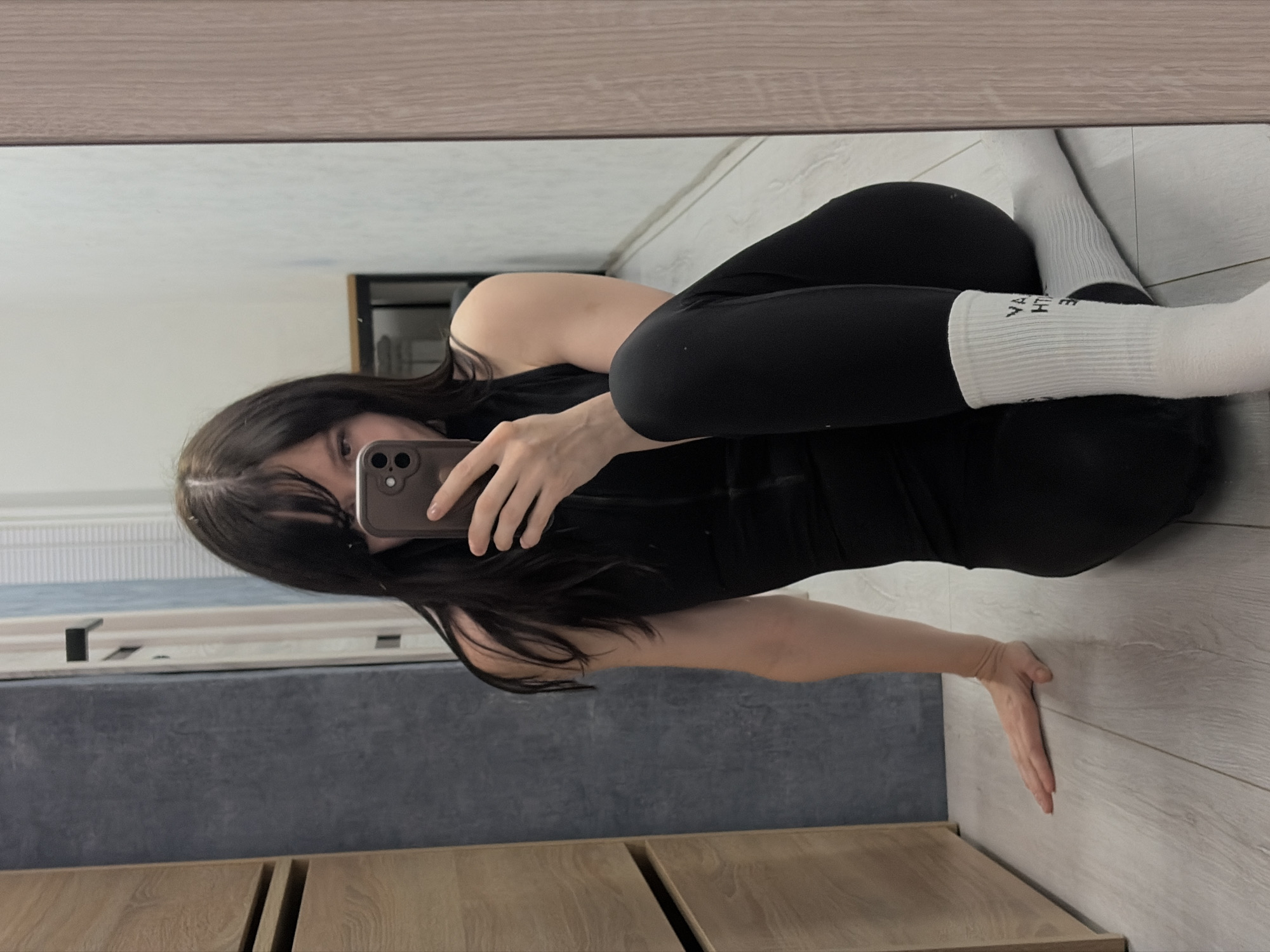 The image is a mirror selfie taken in an indoor setting, possibly an apartment or house, located in what appears to be the city of Chaikovsky, Russia, based on certain clues. The subject is a young woman with dark hair wearing black athletic leggings and a black top, along with white socks that have text on them. She is holding a smartphone in her right hand, presumably taking the picture. The background shows interior elements, including a wood-paneled wall, a grey wall or furniture surface, and light-colored flooring. The lighting is diffused, suggesting it might be daytime. The reflection reveals a glimpse of a window frame. The composition is slightly angled, possibly to capture a better view of the reflection.