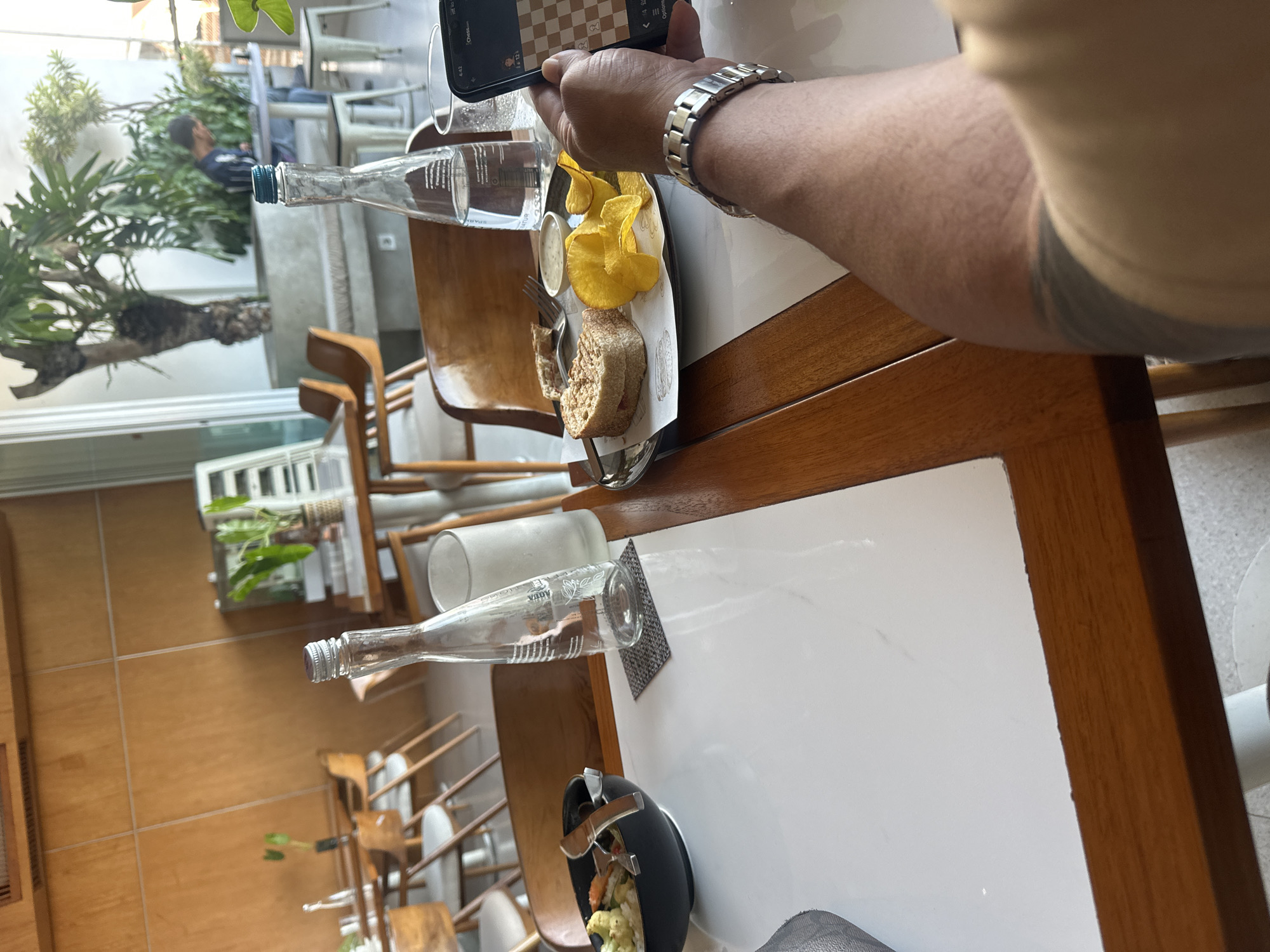 A person's arm and wrist wearing a silver watch are in the foreground, holding a smartphone displaying a chess game. In the background, a white table holds two bottles of water, one clear and one partially filled, a glass, and a plate with a sandwich and potato chips. To the left, a black bowl contains food, and a fork and knife are placed on the table. The restaurant setting features wooden furniture and large windows, with greenery visible outside. The image is taken in a restaurant in North Kuta, Indonesia.