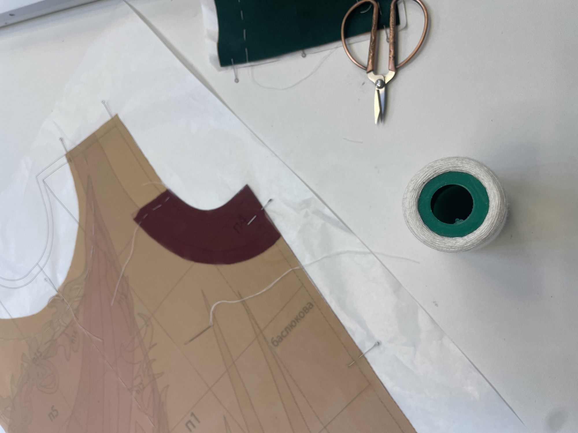 A close-up shot shows a sewing project laid out on a white surface, likely a table, in Pargolovo, Russia. A sewing pattern, printed on translucent paper, is layered with pieces of fabric. One piece is a curved section of deep red fabric, and another is a dark green fabric piece. Pins are used to hold the pattern and fabric together. A spool of white thread is positioned in the lower center of the frame, next to a pair of small, ornate scissors with copper-colored handles. The background is out of focus, but appears to be a flat, neutral surface. The scene depicts an ongoing sewing or tailoring activity.