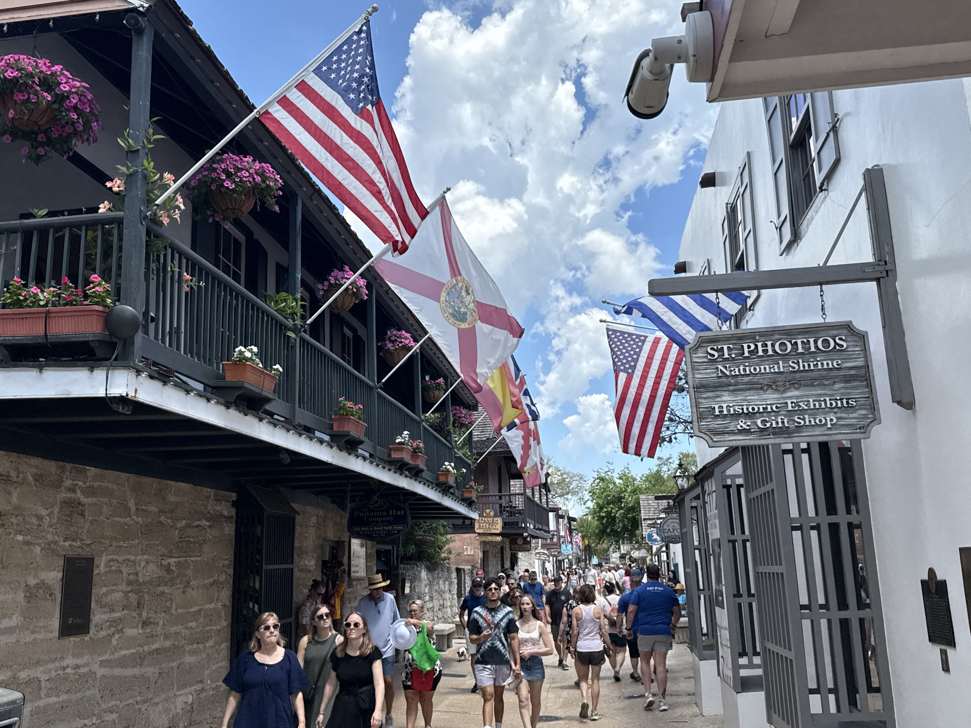 The image captures a bustling street scene in St. Augustine, Florida, with pedestrians strolling along a narrow road flanked by historic buildings. American and Florida flags, along with other flags, are prominently displayed above the buildings. Flower baskets and planters adorn the balconies and storefronts, adding a touch of color to the scene. On the right side of the image, a sign for "St. Photios National Shrine Historic Exhibits & Gift Shop" hangs from a building with white walls and black shutters. A security camera is visible above the sign. The buildings on the left appear to be constructed of weathered stone. The sky is partly cloudy. The presence of signs for "Panama Hat Company" and "Spanish Bakery" further identifies the area as a tourist destination.