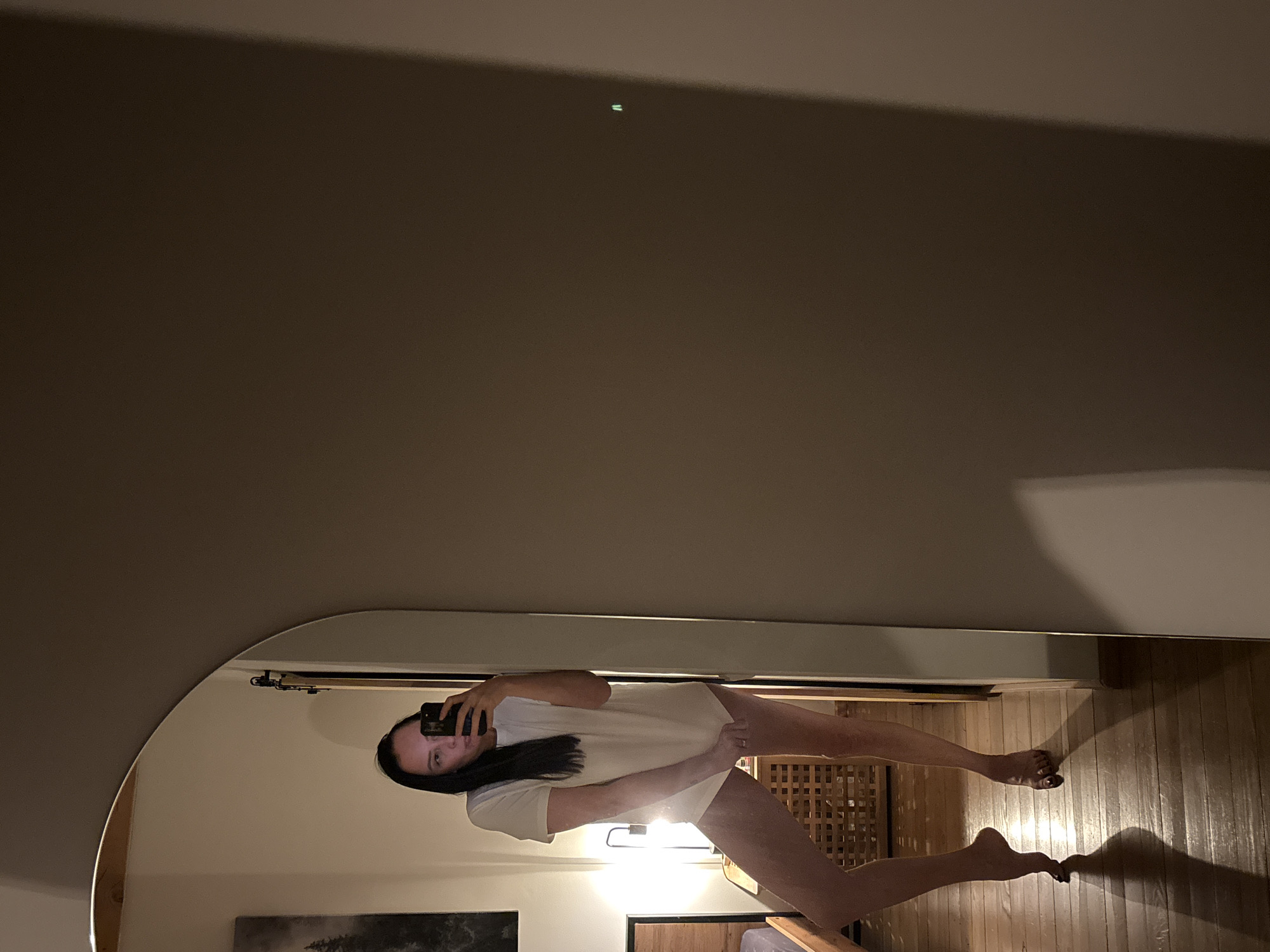 A woman is visible in a mirror, taking a selfie with a smartphone. She's wearing a white t-shirt and appears to be barefoot. The room behind her has wooden flooring, a wooden lattice panel, and a bed. The reflection suggests an interior setting, possibly a bedroom or living space. No specific details indicate that the image was taken in Chaikovsky, Russia.