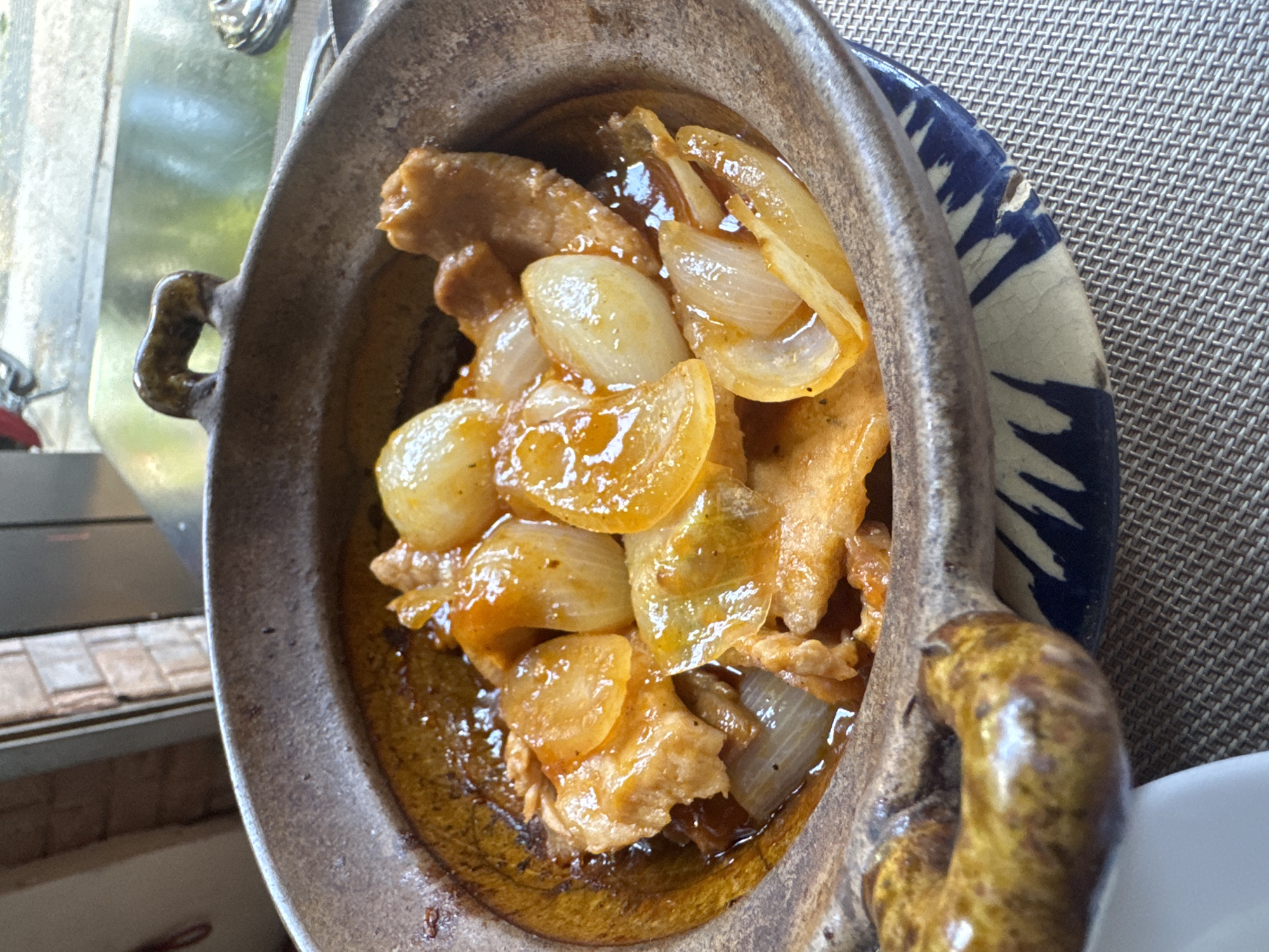 The image presents a top-down view of a savory dish served in a rustic, oval-shaped clay pot. The pot contains what appears to be chunks of meat, possibly chicken or pork, braised or stewed with numerous large, translucent, and caramelized onion pieces. The ingredients are coated in a thick, glossy, golden-brown sauce. The interior of the clay pot has a smooth, dark orange-brown glaze, contrasting with its unglazed, textured, grayish-brown exterior and small, glazed loop handles.
The clay pot is set upon a round ceramic plate, which features a distinctive blue and white pattern, resembling abstract brushstrokes or traditional motifs, around its rim. A small chip is visible on the blue-patterned edge of this plate. Beneath the plate is a light brown or beige placemat with a finely woven, grid-like texture.
The setting appears to be a dining area during the daytime, suggested by the bright, natural light reflecting off a blurred window or glass surface in the background to the left. Below this reflection, a tiled surface with small, rectangular tiles is partially visible. To the right of the main dish, a portion of a plain white ceramic object, possibly another plate or bowl, is discernible.
The scene captures a prepared meal, highlighting its appetizing appearance and the traditional serving style. No individuals are present, and no visible text is discernible in the media file.