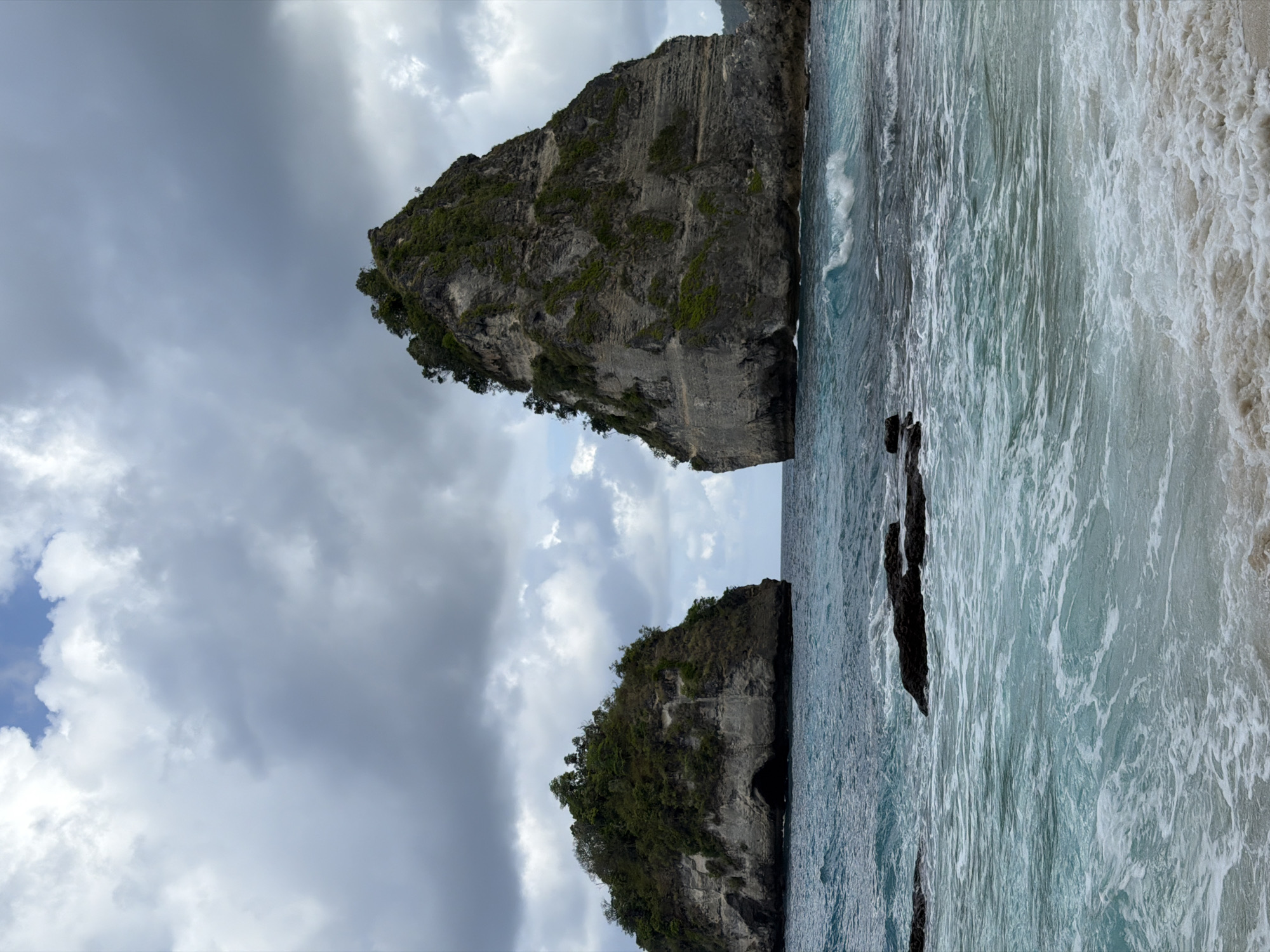 Two large, rocky islands topped with dense green foliage rise from a turbulent turquoise ocean under a cloudy sky. The island on the right is significantly larger and steeper than the one on the left, which has a visible cave opening at its base. The foreground shows crashing waves with white foam moving towards a sandy shore. Black rocks are scattered in the shallow water between the shore and the islands. The scene captures the dramatic natural beauty of a coastal area, likely a beach or bay, on Nusa Penida, Indonesia.