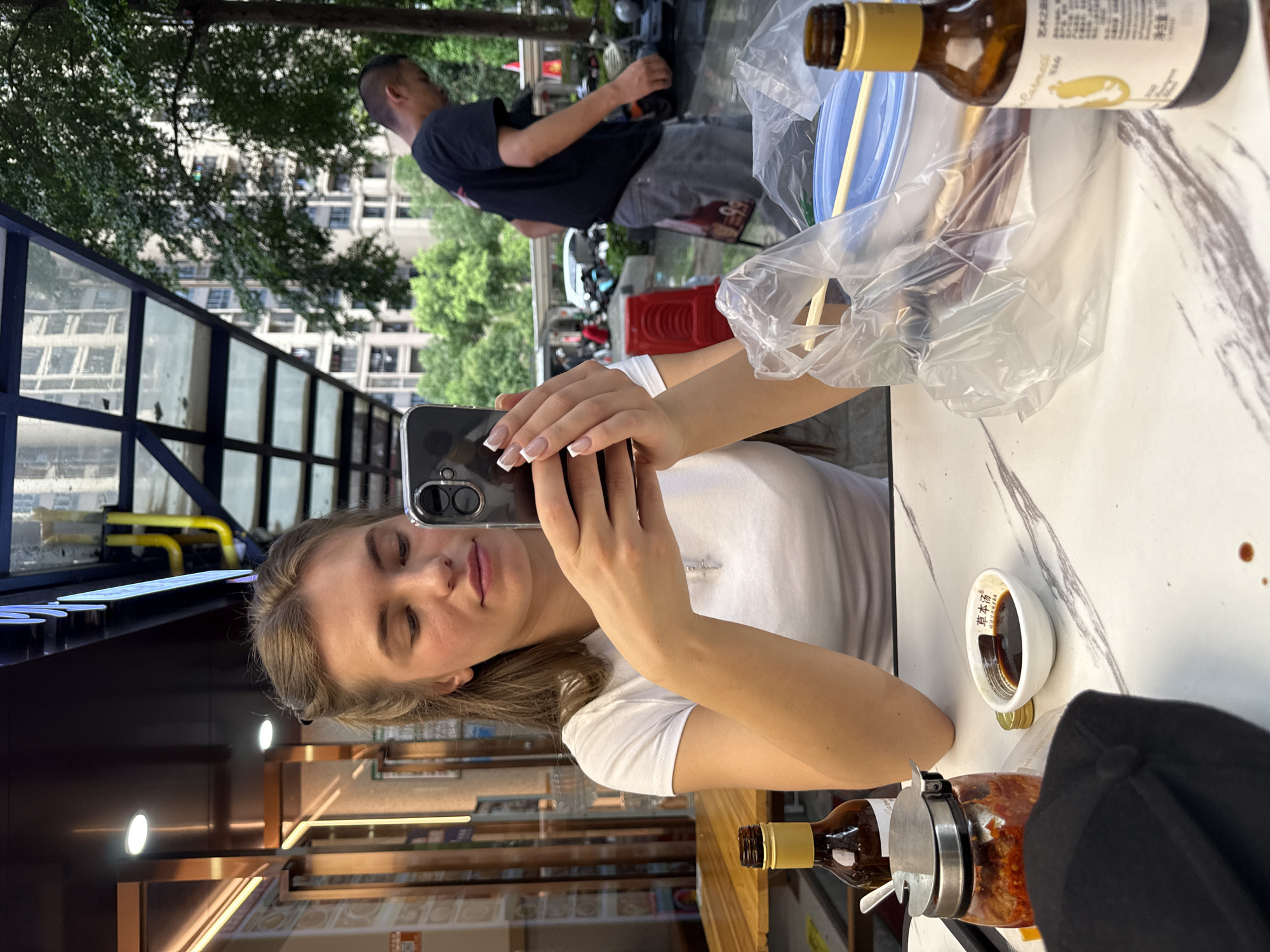 A young woman with blonde hair, wearing a white t-shirt, takes a selfie with her phone. Her left hand holds the phone, and her right arm is visible, with a silver necklace with a cross pendant. She is seated at a table with a marble-like surface, where a glass bottle with a yellow cap, a clear plastic bag with chopsticks, and a small white bowl with dark liquid are placed. In the background, a man in a dark t-shirt and grey pants stands near a motorcycle, with trees and buildings visible. The scene appears to be outdoors, possibly a street-side dining area, with an urban setting. The lighting suggests it is daytime. The woman's expression is neutral as she takes the photo. There is some Chinese text on the label of the bottle, and also on the small white bowl, which reads "酱车汤". The image is captured from a slightly elevated angle, looking down on the woman.