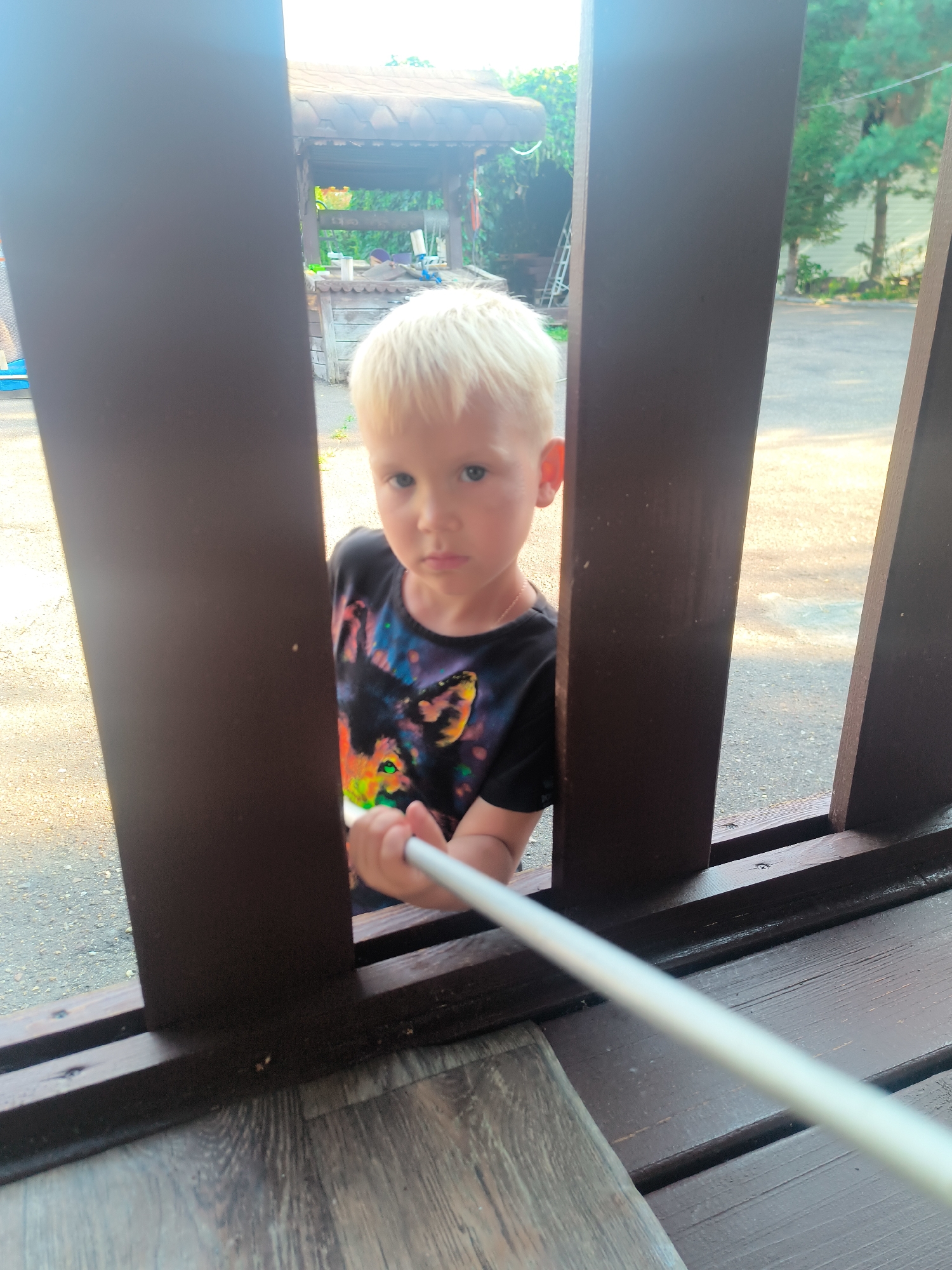 A young boy with blond hair peers through wooden slats, his gaze directed forward. He is wearing a black t-shirt with a colorful, abstract design. His right hand is extended, holding a white cylindrical object, possibly a stick or a pole. The background suggests an outdoor setting, with trees and a structure that resembles a well or a gazebo visible. The ground appears to be asphalt or a similar paved surface. The overall impression is a candid moment of a child engaged in an activity outdoors.