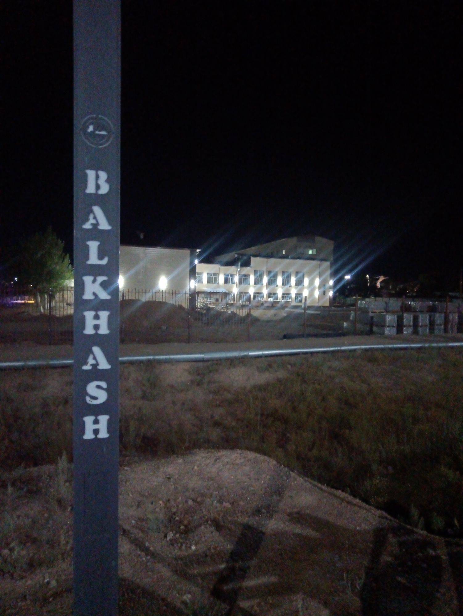 A vertical, dark gray pole stands in the foreground, painted with white block letters vertically arranged. The letters spell "BALKHASH". At the top of the pole, a circular symbol with a cigarette and a line through it is visible. Behind the pole, a low fence runs horizontally. Beyond the fence, a two-story building with many lit windows is visible, suggesting it is nighttime. The building appears to be a school or an administrative facility, with a light-colored facade. To the right of the building, stacks of what appear to be building materials, possibly concrete blocks, are piled up. The ground in front of the building and around the stacks of materials is uneven and appears to be unpaved. A tree with green foliage is situated to the left of the building. The overall scene depicts an outdoor area at night, possibly on the outskirts of Balqaş, Kazakhstan, given the inscription on the pole.