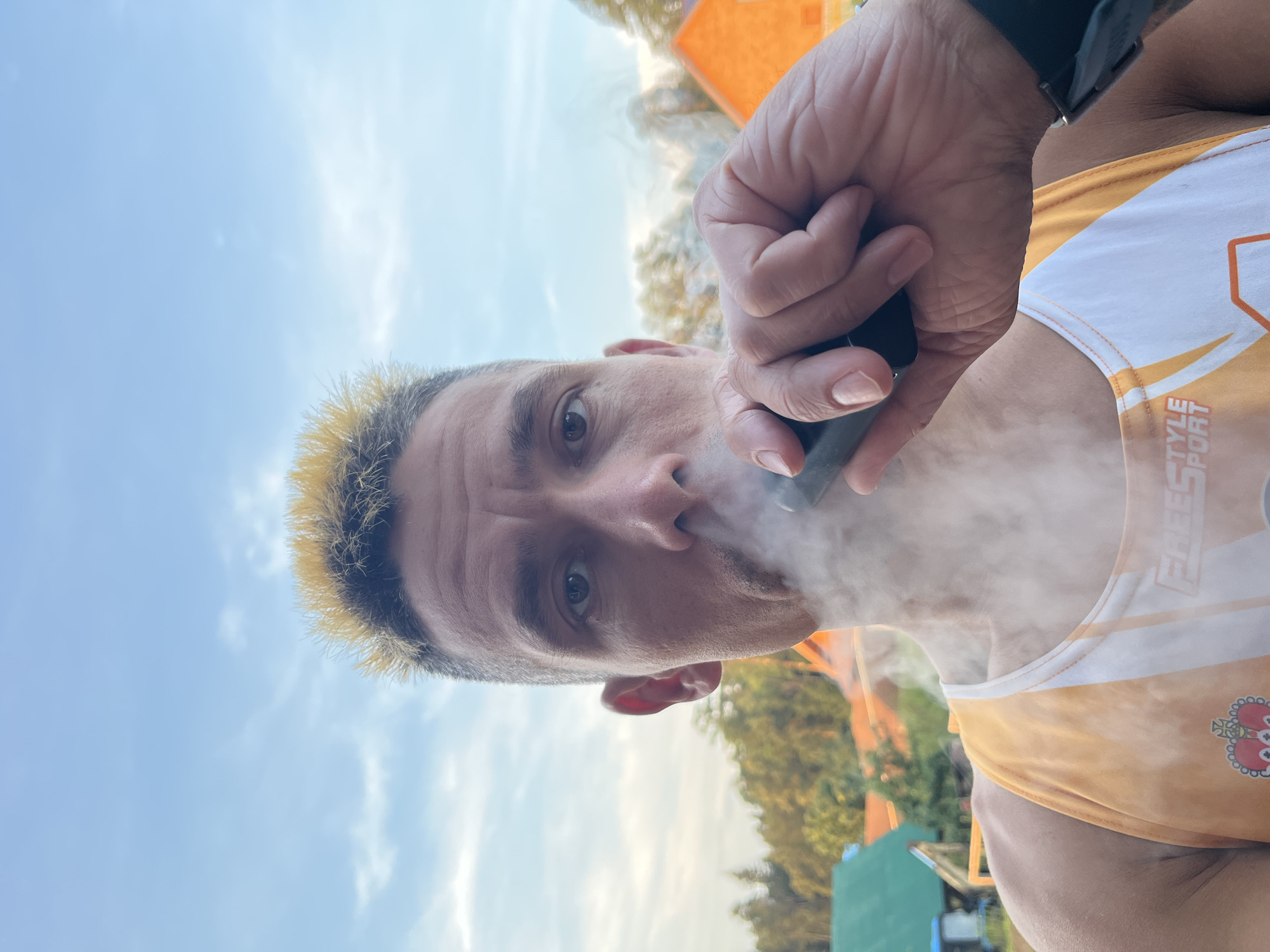 A man with spiky, bleached blonde hair is shown from the chest up, holding a black vape device to his mouth and exhaling a cloud of vapor. He is wearing an orange and white tank top with the text "FREESTYLE SPORT" and a crown emblem. The background consists of trees, houses, and a pale blue sky with scattered clouds. The image appears to be taken outdoors, possibly in a residential area. The man's facial expression is neutral, and his eyes are open.