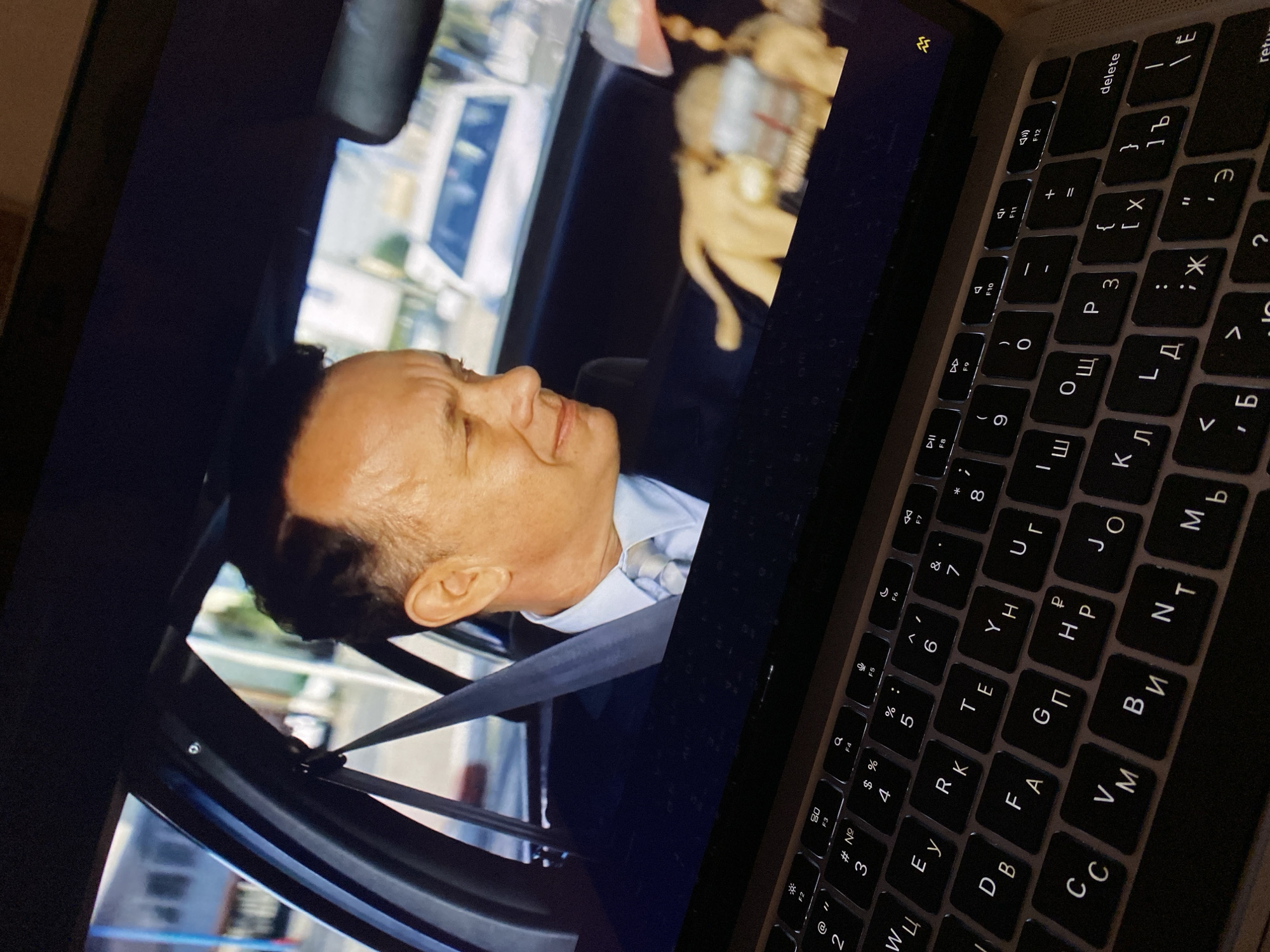 In this image, a laptop screen displays a scene from a movie, featuring a man, possibly Tom Hanks, in the driver's seat of a car, wearing a suit and tie. The movie depicts him looking towards the right side of the frame, with a blurry background suggesting an urban setting. In the foreground, a laptop keyboard with Cyrillic characters is visible, indicating a close-up view of someone using the device to watch the movie. The lighting suggests the room is dimly lit, with the screen and keyboard backlight being the primary light sources. The specific location of Kuta Selatan, Indonesia, is not discernible from the provided image.