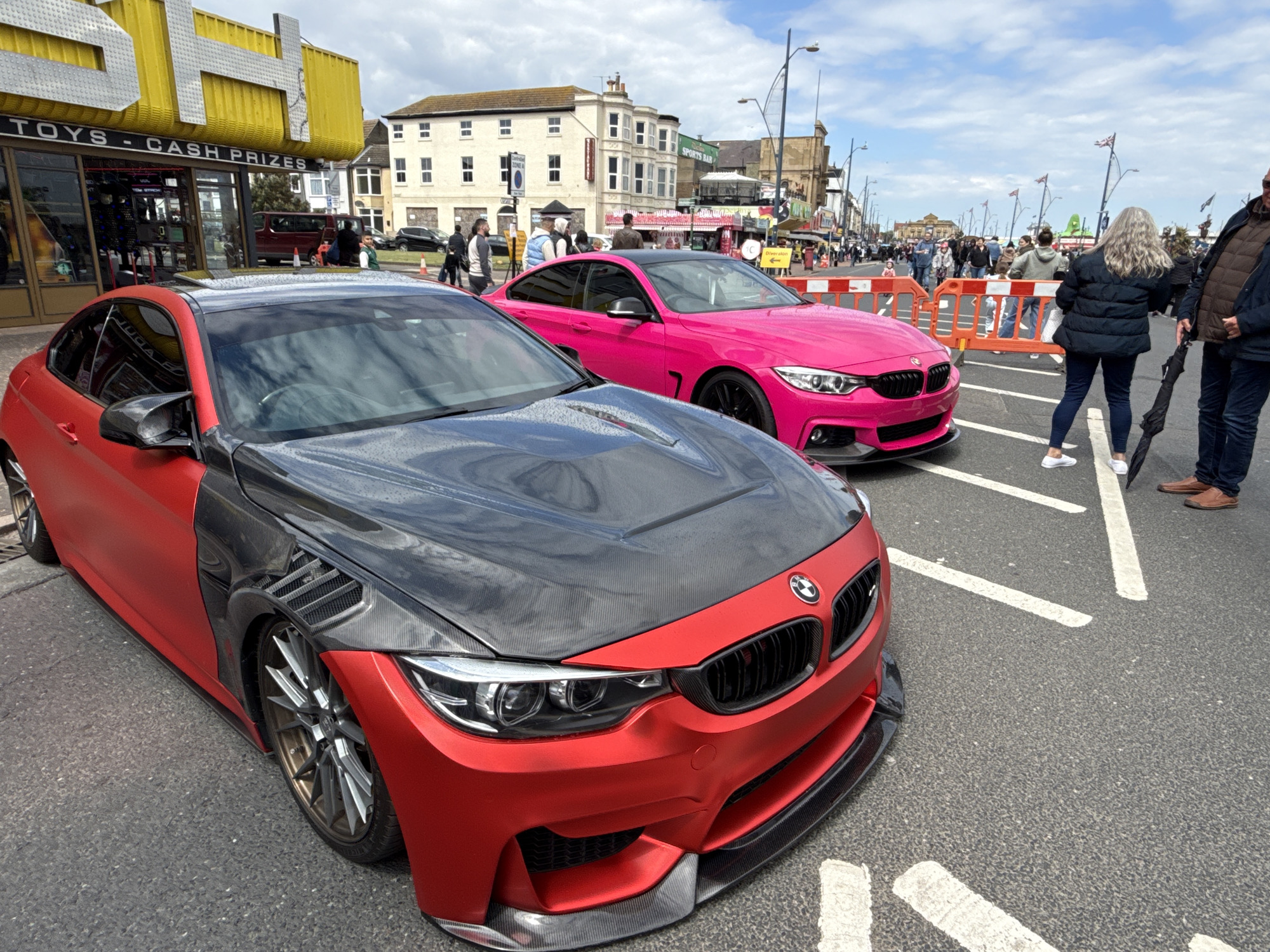 The image shows a street scene in Great Yarmouth, United Kingdom. Two customized BMW cars are parked on the street, which is also being used as a pedestrian thoroughfare. The car in the foreground is a red-and-black custom model, featuring a black hood and carbon fiber detailing on the side panels. Behind it, a pink BMW stands out against the backdrop. Pedestrians, some partially visible, are walking along the street, separated from the cars by temporary orange barriers. Shops with yellow awnings and signs reading "TOYS - CASH PRIZES" line the street on the left. The architecture in the background includes buildings with white facades and multiple windows. Union Jack flags adorn the streetlights. The sky is partly cloudy.