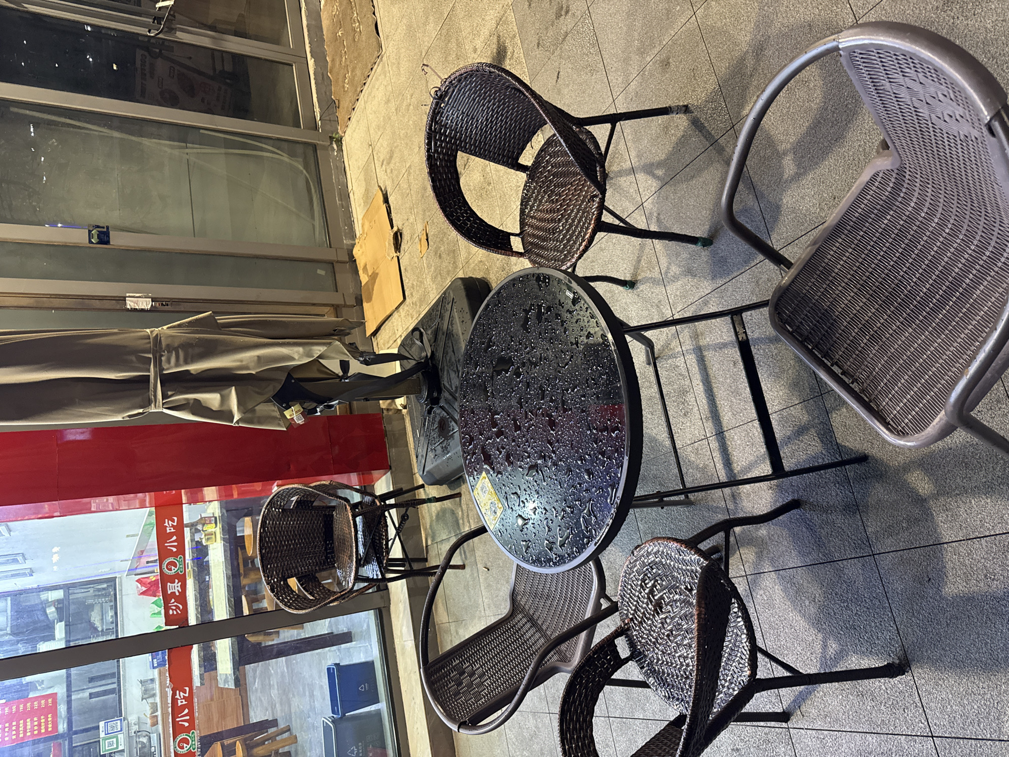 This image depicts an outdoor seating area in front of a business located on Nanjing Road Pedestrian Street in the Huangpu District of Shanghai, China. A small, round, dark-colored table with visible water droplets sits in the center of the frame, suggesting recent rain. Four woven chairs surround the table. Two chairs are dark brown rattan, and two appear to be a lighter brown rattan. A fifth, metallic gray chair is partially visible in the lower right corner of the image. Behind the table and chairs, a large, light brown umbrella is folded and tied up. Through the glass windows of the establishment, tables and chairs are visible inside, along with signage in Chinese. Two blue recycling bins are positioned near the window to the left. The ground is paved with light gray tiles, and shadows are cast by the table and chairs. The overall scene suggests a temporarily closed outdoor dining or waiting area.