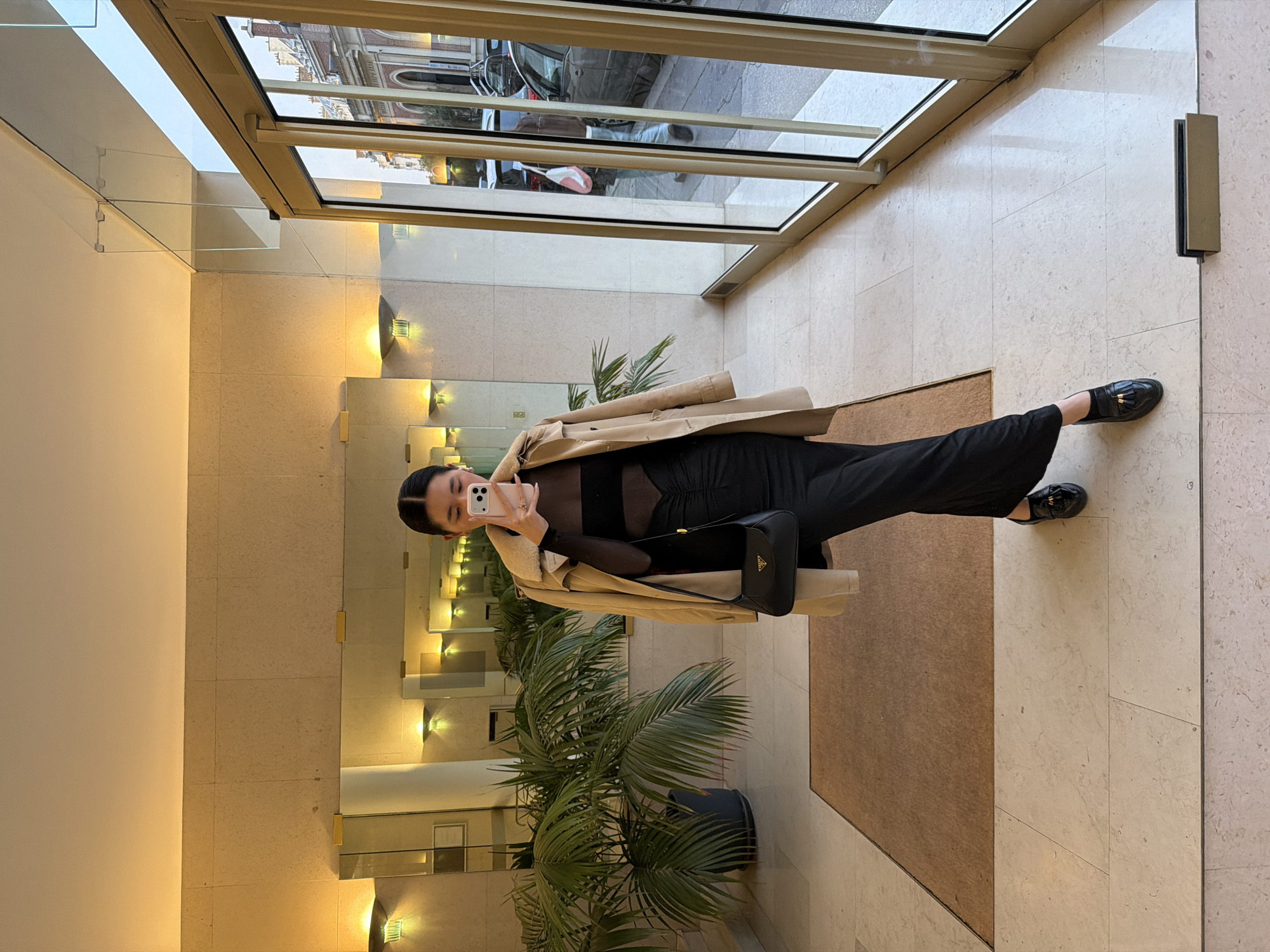 A woman takes a selfie in what appears to be the entrance lobby of a building. She is wearing a black outfit, a beige trench coat, and black loafers. She is carrying a black handbag. The building's entrance doors are glass and offer a view of an exterior Parisian street with cars and buildings visible. The lobby has light-colored marble flooring and wall panels, mirrors, wall-mounted lights, and potted plants.