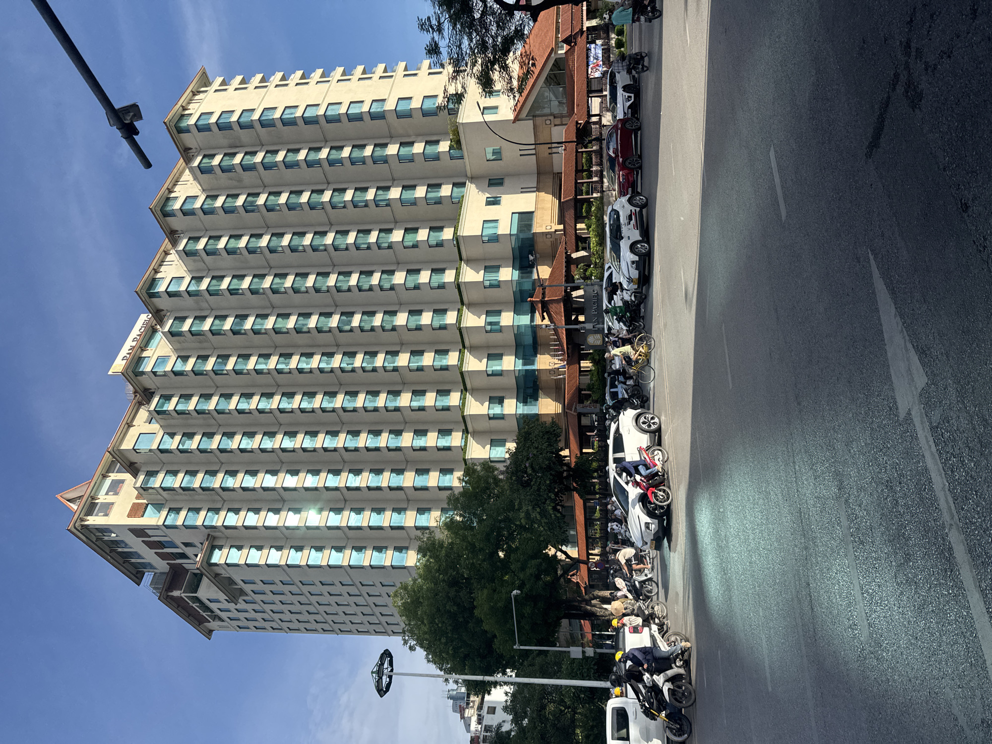 This is a medium shot, outdoor, eye-level, daytime, horizontal photograph of a busy intersection in front of a large, multi-story hotel building. The building, identified by the "PAN PACIFIC" sign on its roof, is light-colored with numerous blue-tinted windows and a reddish-brown tiled roofline. Trees with green foliage are visible to the left of the building and also in the mid-ground. The foreground shows a paved road with several vehicles and people.
In the street, there are many motorbikes, some carrying one or two people. People are wearing helmets, and some are dressed in casual clothing. Several cars are also present, including a white SUV, a red sedan, and another white car. A white truck is parked on the left side of the frame. A lamppost with a circular light fixture is visible on the left. The sky is clear and blue. The overall scene depicts a typical urban environment with significant traffic and pedestrian activity. The architecture of the hotel suggests it is a modern establishment in a developed city. Given the name "Pan Pacific" and the architecture, this location is likely the Pan Pacific Hanoi hotel, situated in the city of Hanoi, Vietnam.