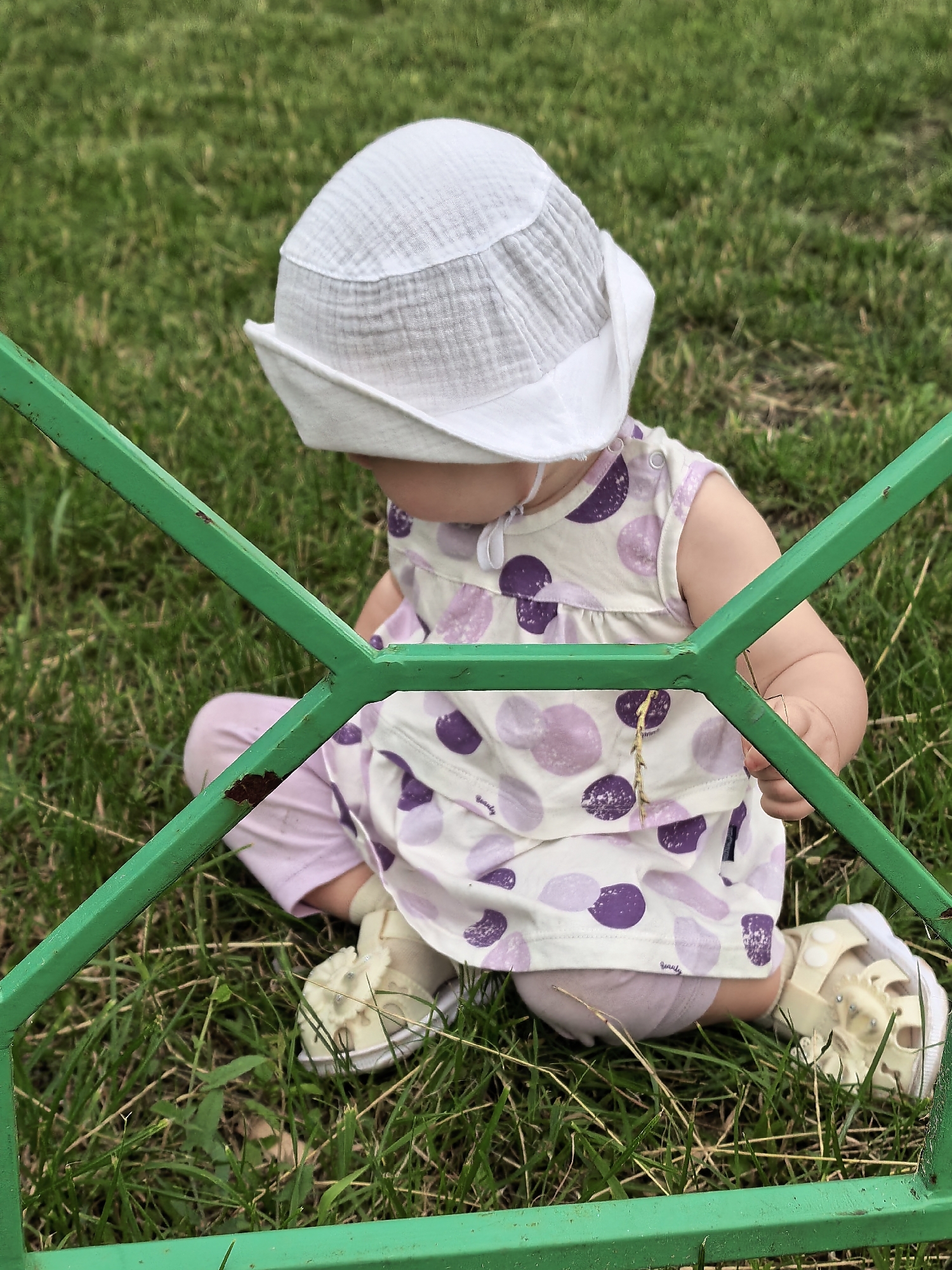 A baby wearing a white sun hat and a light-colored dress with purple polka dots sits on the grass behind a green metal fence. The baby's legs are bent, and its feet are in light-colored shoes. The grass appears to be a lawn. The scene is outdoors, with no other people or objects clearly visible. The location is not specified in the image, but the request specifies Sterlitamak, Russia.