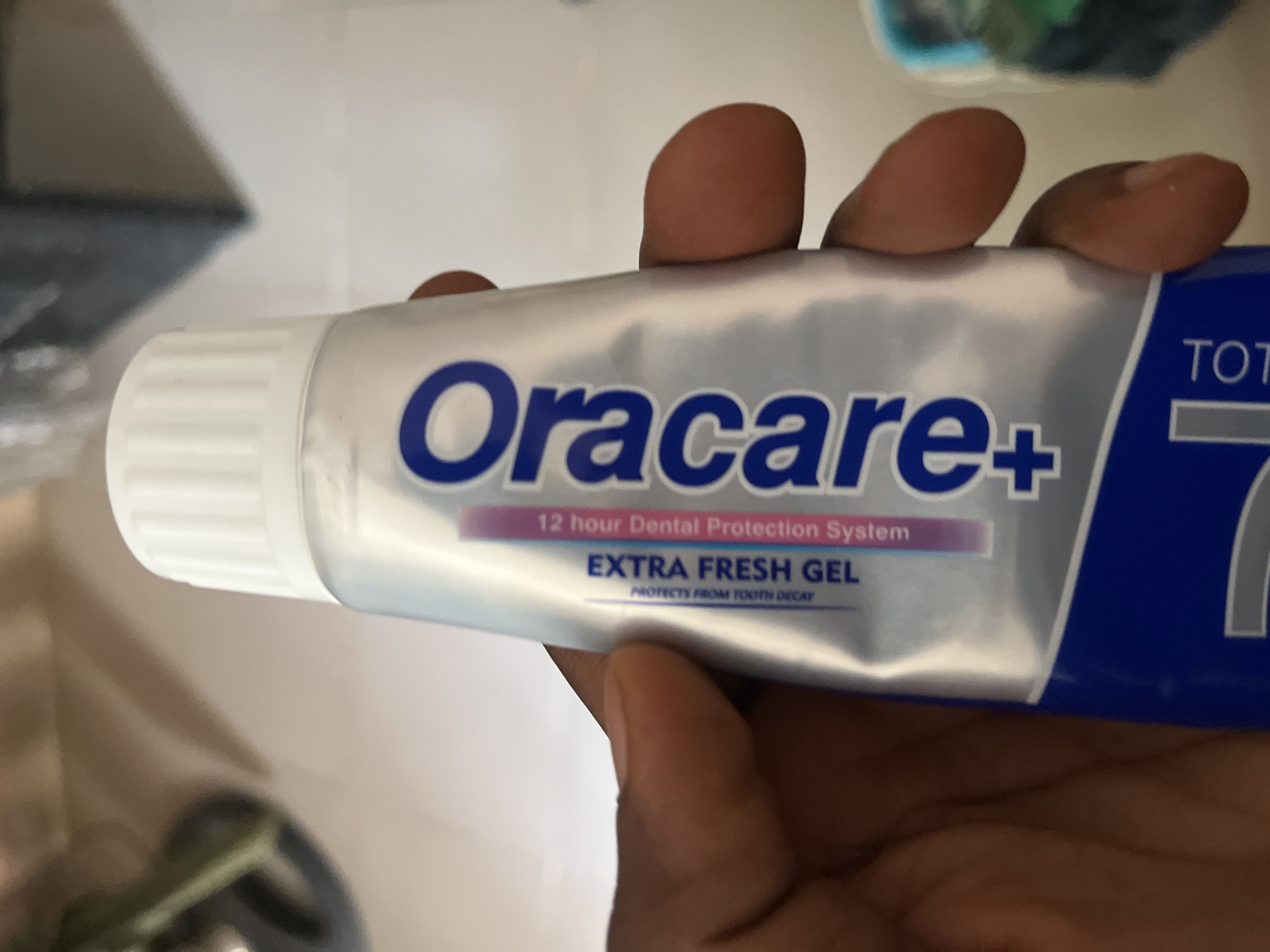 A tube of Oracare+ Extra Fresh Gel toothpaste is held by a person's hand. The toothpaste tube is silver with blue and pink accents and features the brand name "Oracare+" in large blue letters. Text on the tube reads "12 hour Dental Protection System" and "EXTRA FRESH GEL PROTECTS FROM TOOTH DECAY". The image is taken from a slightly elevated angle, and the background is out of focus, suggesting an indoor setting. No specific location in Jalingo, Nigeria, can be identified from the image.
