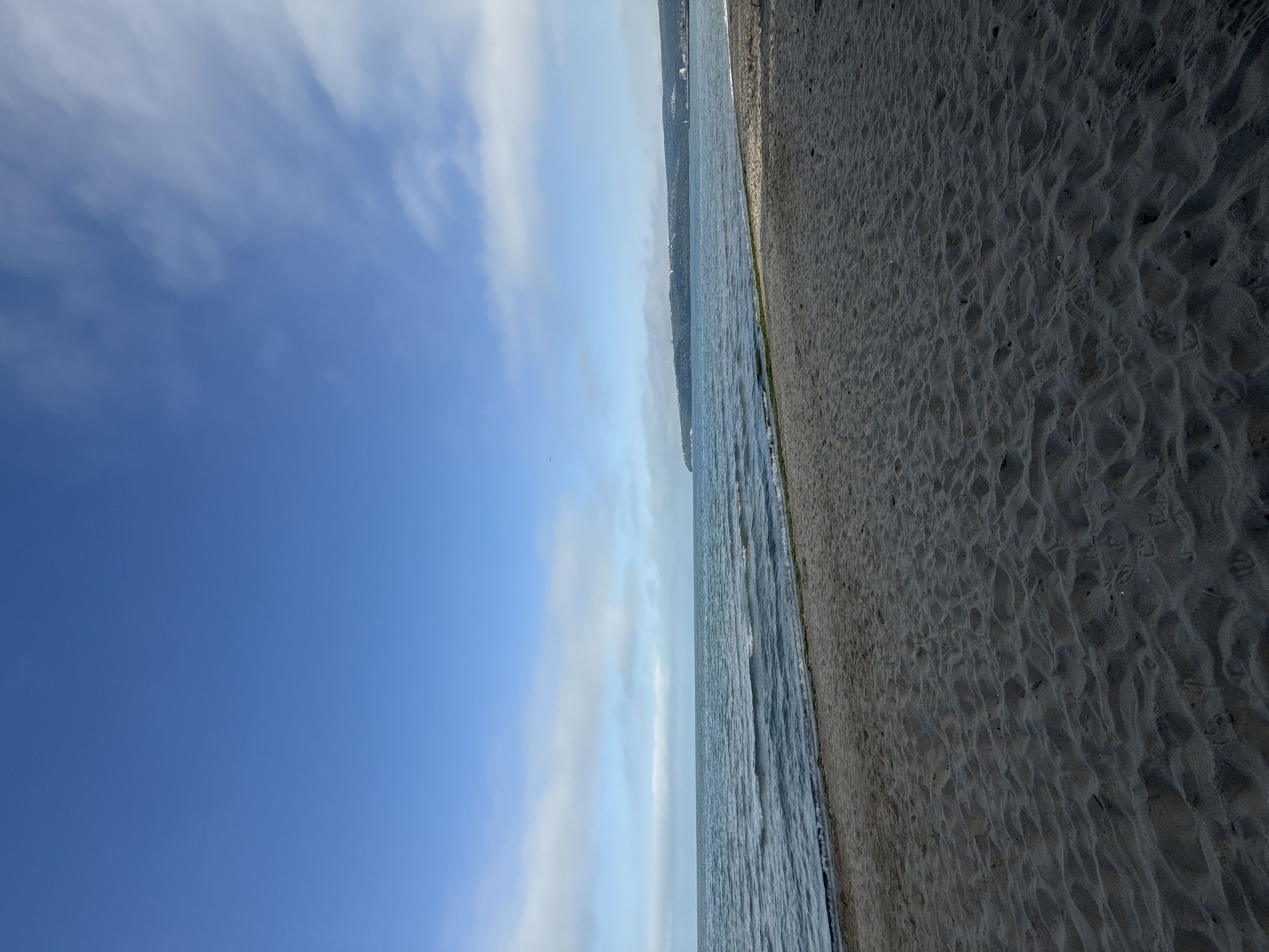 The image depicts a beach scene in Varna, Bulgaria. The composition is divided roughly into thirds: the sky on the left, the sea in the middle, and the sandy beach on the right. The sky is blue with some wispy clouds. The sea is a darker blue-grey, with visible waves. The sand is a dark grey-brown. There are no people visible in the image, but the overall view suggest the city is on the other side of the sea.
