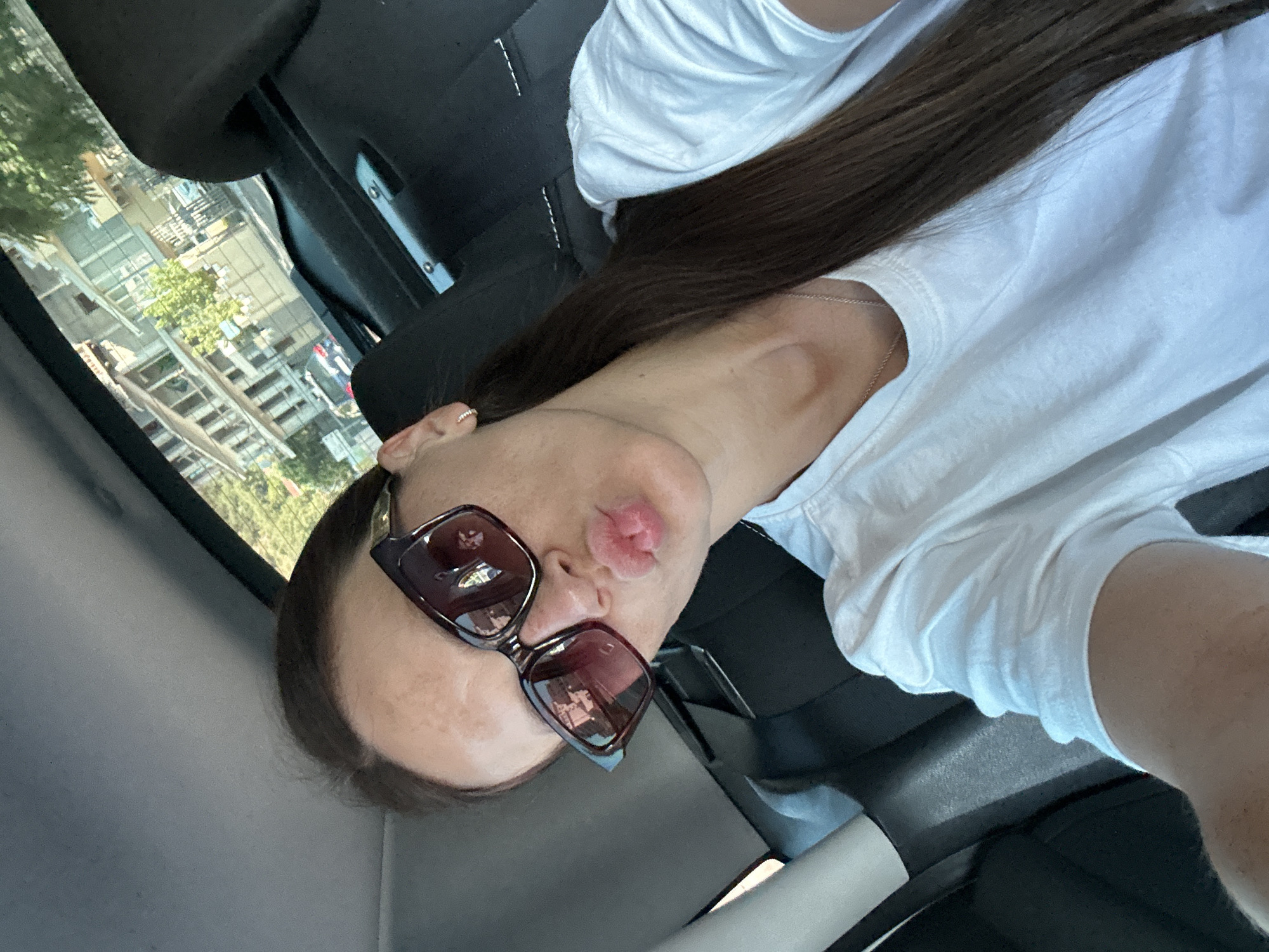 A woman is in the back seat of a car, facing the camera and making a kissing gesture with her lips. She is wearing large, dark-rimmed sunglasses, a white t-shirt, and a thin silver necklace. Her long, dark hair is pulled back into a ponytail. The interior of the car is visible, with black upholstery and some car features. Through the car window, a multi-story building with balconies and trees can be seen, suggesting an urban environment, possibly in Budva, Montenegro, given the context. The lighting indicates it is daytime.