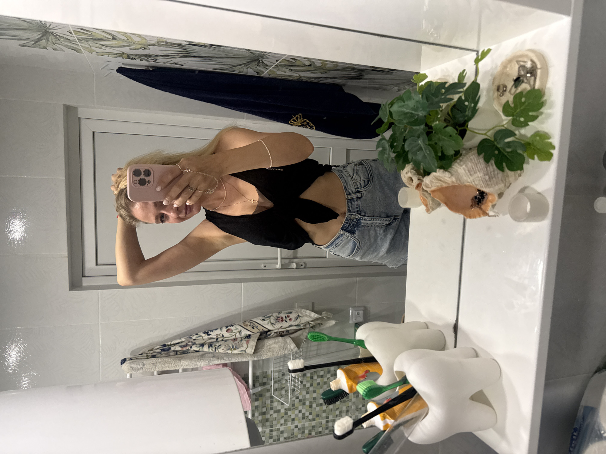 A blonde woman with her hand in her hair is taking a mirror selfie in a bathroom. She is wearing a black, tie-front crop top and denim shorts. She has several rings on her fingers and a necklace with a cross pendant. In the foreground, there are two white, tooth-shaped toothbrush holders with toothbrushes and toothpaste inside. To the right of the toothbrush holders, there is a shell with a plant and two small white votive candle holders. The bathroom has white tiled walls and a white door.