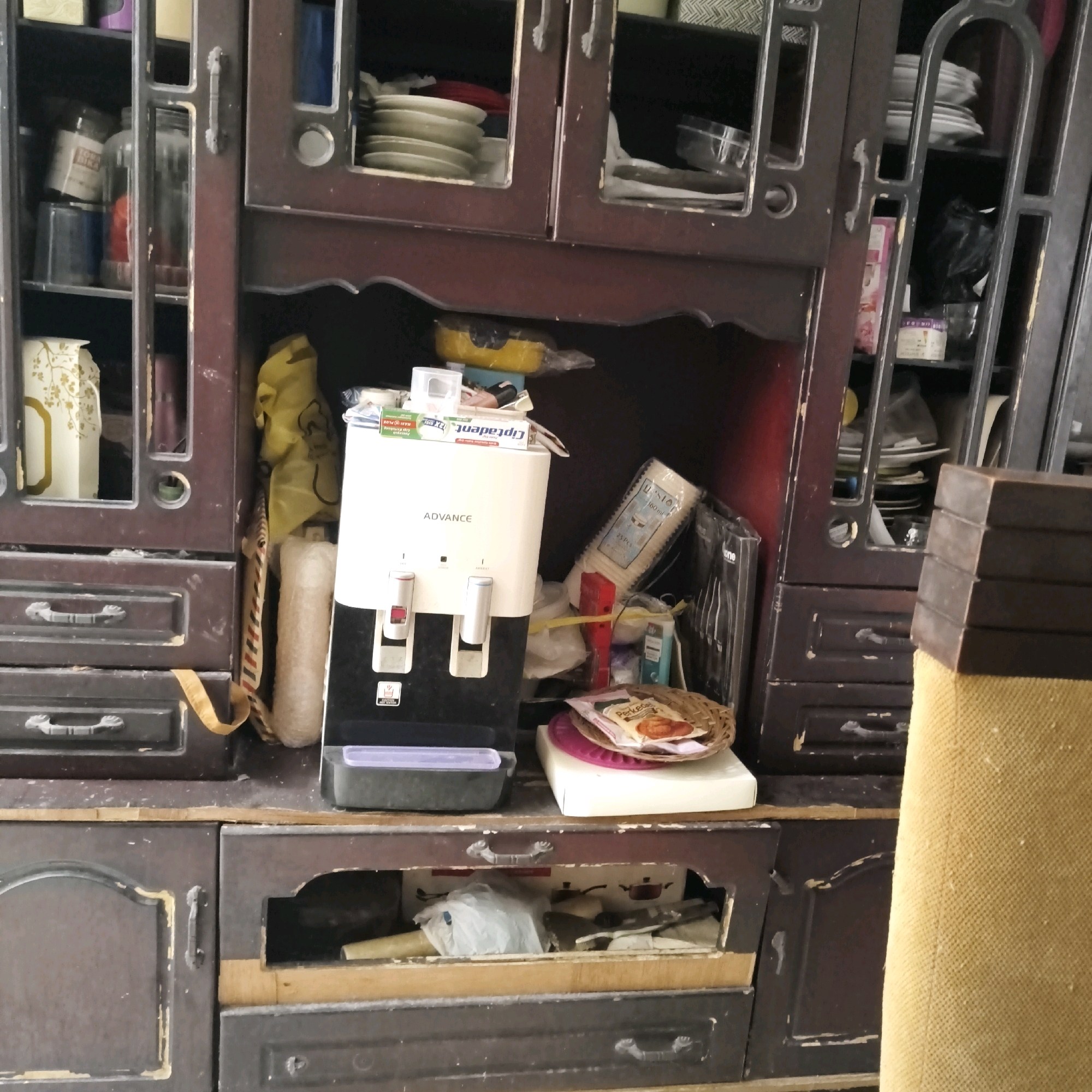 The image depicts a cluttered antique wooden cabinet in what appears to be a residential setting in South Jakarta, Indonesia. The cabinet is dark brown with some visible signs of wear and tear, including chipped paint and faded wood. It contains various items on its shelves and within its drawers.
On the top shelf, several stacks of white plates are visible behind a glass-paned door. To the right of the plates, a black plastic bag is partially visible. Below this, a white water dispenser with a black base is the most prominent object in the center of the image. It has two spigots, one red and one blue, and a brand name "ADVANCE" printed on its front. Above the water dispenser, a tube of toothpaste, labeled in a foreign language, is placed on top of the dispenser. To the right of the dispenser, a stack of white plastic cups is leaning against a dark surface. A small woven basket containing food items is placed on a white rectangular object to the right of the water dispenser.
The lower part of the cabinet features several drawers with ornate handles. One drawer is partially open, revealing plastic bags and other unidentifiable objects inside. To the far right of the image, a portion of a textured yellow object, possibly a piece of furniture or fabric, is visible. The overall impression is one of a lived-in space with items stored in a somewhat disorganized manner. The lighting suggests it is daytime, possibly indoors with natural light filtering in.