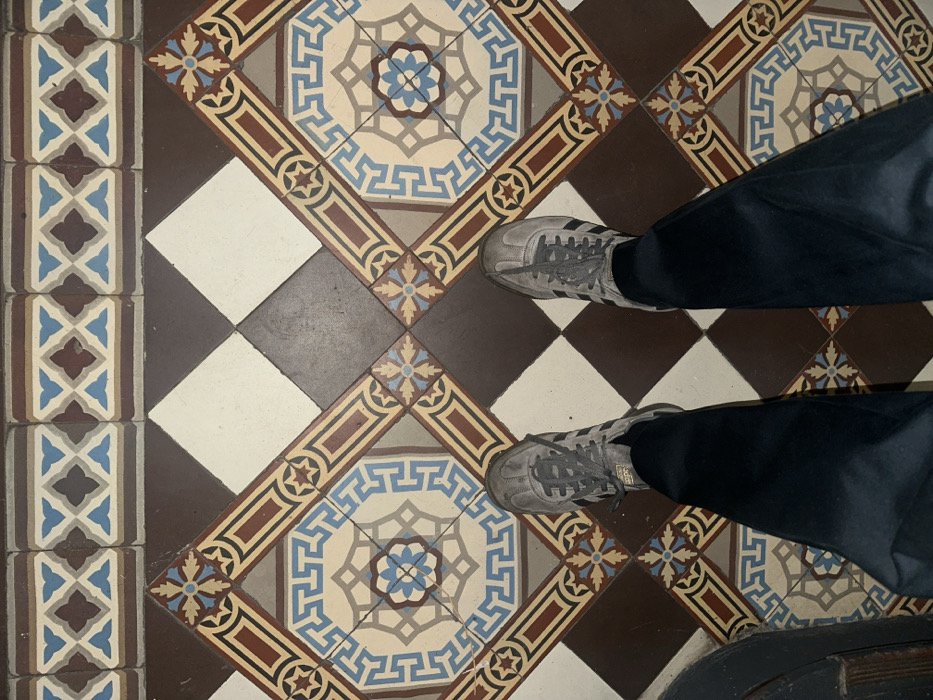 A pair of feet, clad in dark blue trousers and grey, worn sneakers with dark laces and three side stripes, stands on an intricately patterned tiled floor in an interior setting in Carnin, France. The floor features a main field of diagonally set square tiles, alternating between solid dark brown and elaborate decorative patterns. The decorative tiles contain a central light blue floral motif encircled by a light brown Greek key pattern, all framed by red, brown, and cream geometric borders. These are interspersed with smaller square tiles bearing light blue floral designs at their corners. A distinct border runs along the top, composed of rectangular tiles with repeating light blue, dark red, and brown geometric patterns on a cream background. The floor exhibits signs of use.