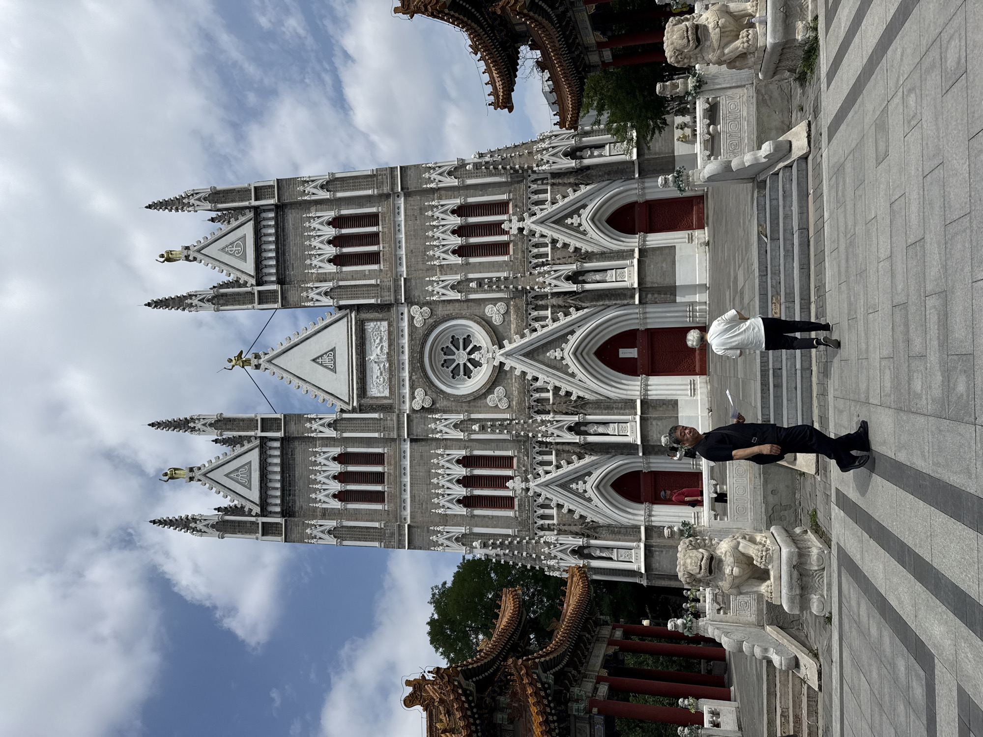 This is a photograph taken outside the North Cathedral, also known as the Church of the Immaculate Conception, located in Xicheng District, China. The cathedral is a grand Gothic-style building with twin spires, ornate stone carvings, and red double doors. In the foreground, two individuals are walking on a paved plaza. To the left and right of the cathedral entrance are traditional Chinese-style pavilions with orange tiled roofs and red pillars. Stone lion statues are positioned on either side of the entrance steps. The sky above is partly cloudy.