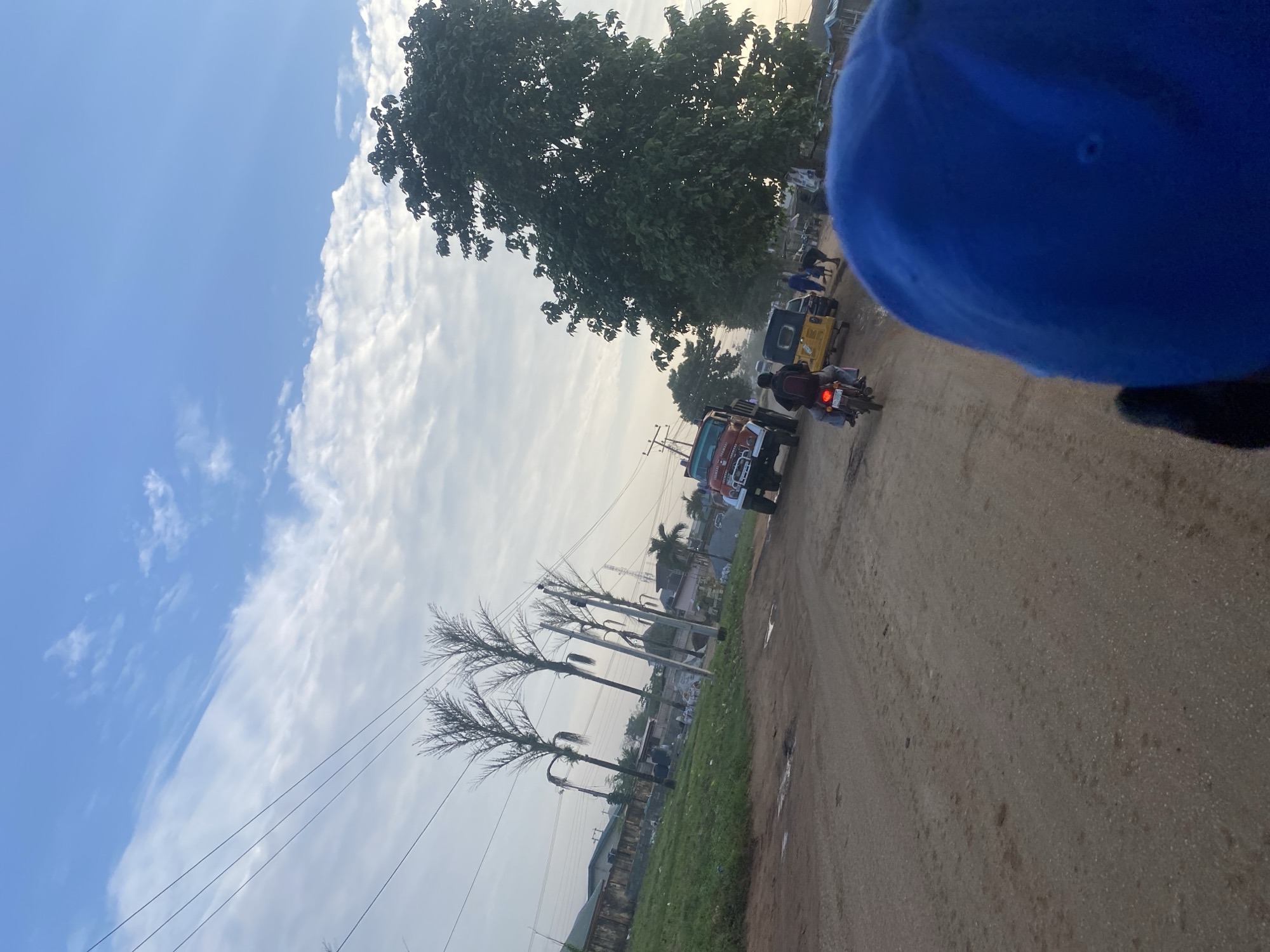 The image depicts a dusty road in Ikorodu, Nigeria, under a sky with scattered clouds. In the foreground, a person wearing a blue cap is partially visible, suggesting the photo is taken from their perspective, possibly on a motorcycle. Further down the road, a motorcycle is being ridden by a person wearing a red shirt and black pants, with a passenger on the back. To the right of the motorcycle, a yellow auto-rickshaw (keke) is traveling, and several pedestrians are present near it. A red truck is parked on the left side of the road, and in the background, buildings and vegetation are visible, including several tall palm trees and a large leafy tree. Electrical poles and wires run across the scene.