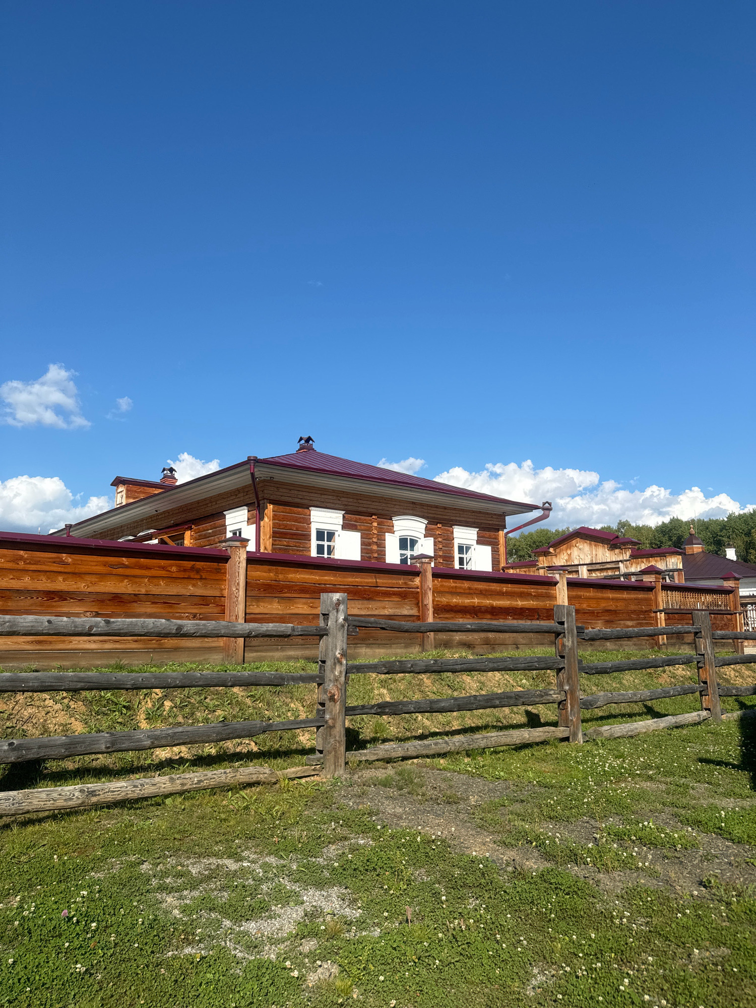 The image depicts a sunny day in the city of Taltsy (Irkutsk), Russia, featuring a traditional wooden house and fence. The house, constructed with logs, has a reddish-brown roof and white-trimmed windows. A wooden fence, made of horizontal logs, separates the property from the foreground, which is covered in green grass and small white flowers. The sky is a clear blue with scattered white clouds. The scene appears peaceful and rustic, characteristic of rural Russian architecture.